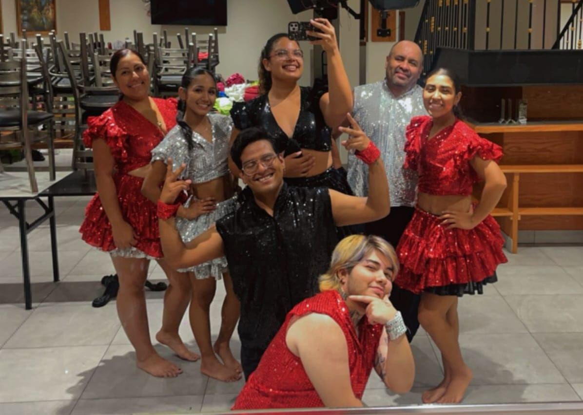 A group of seven people, dressed in sparkly performance outfits, pose for a selfie. The outfits feature red and silver sequins, and they appear to be indoors.