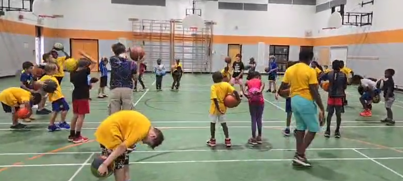 Children practice basketball in a gymnasium. Many hold basketballs and are in various stages of the sport, with a coach observing.