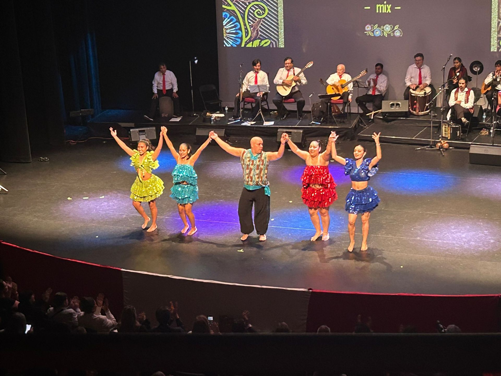 Dancers and a man on stage, holding hands, facing the audience. A band sits behind them.