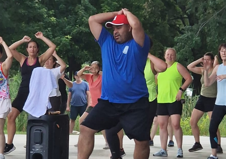 Man in red cap leads outdoor dance class; participants mimic arm movements. Several women in athletic wear are in a park setting.
