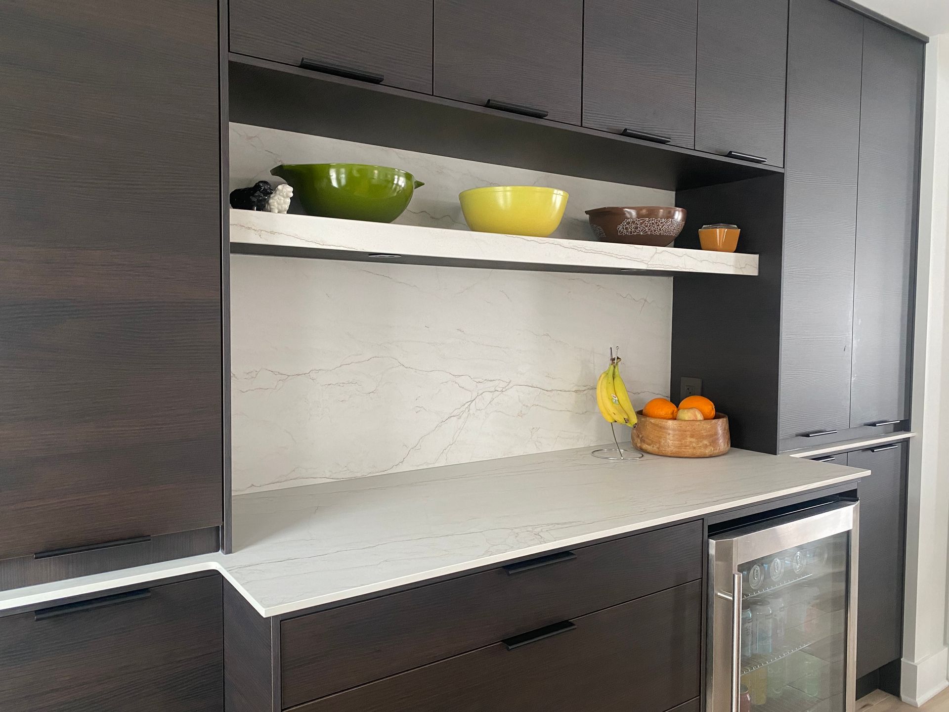 Dark cabinets with pull-down countertop, open shelf with bowls, fruit and beverage cooler.