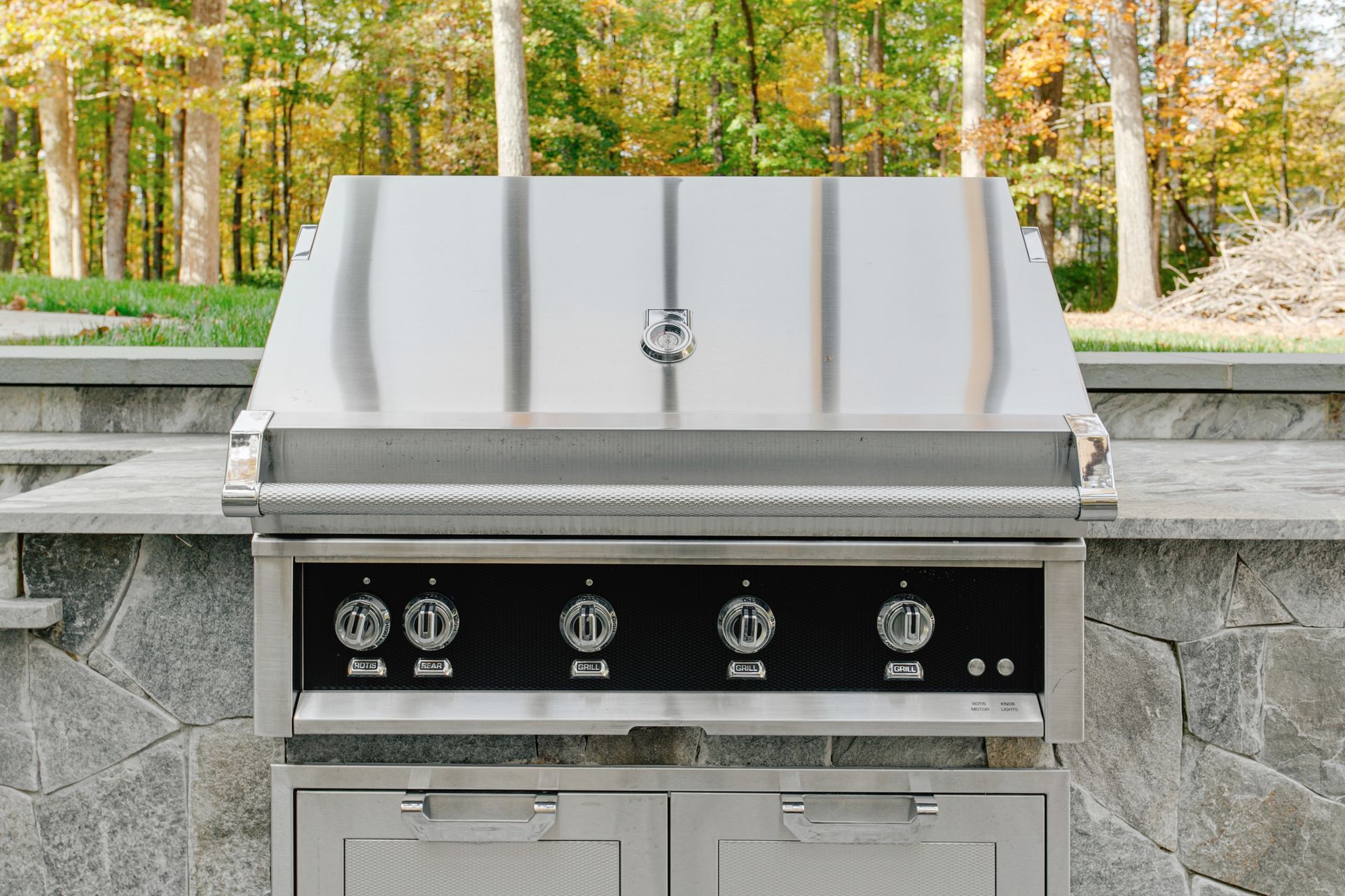 Stainless steel outdoor grill built into stone countertop with knobs and cabinets below.