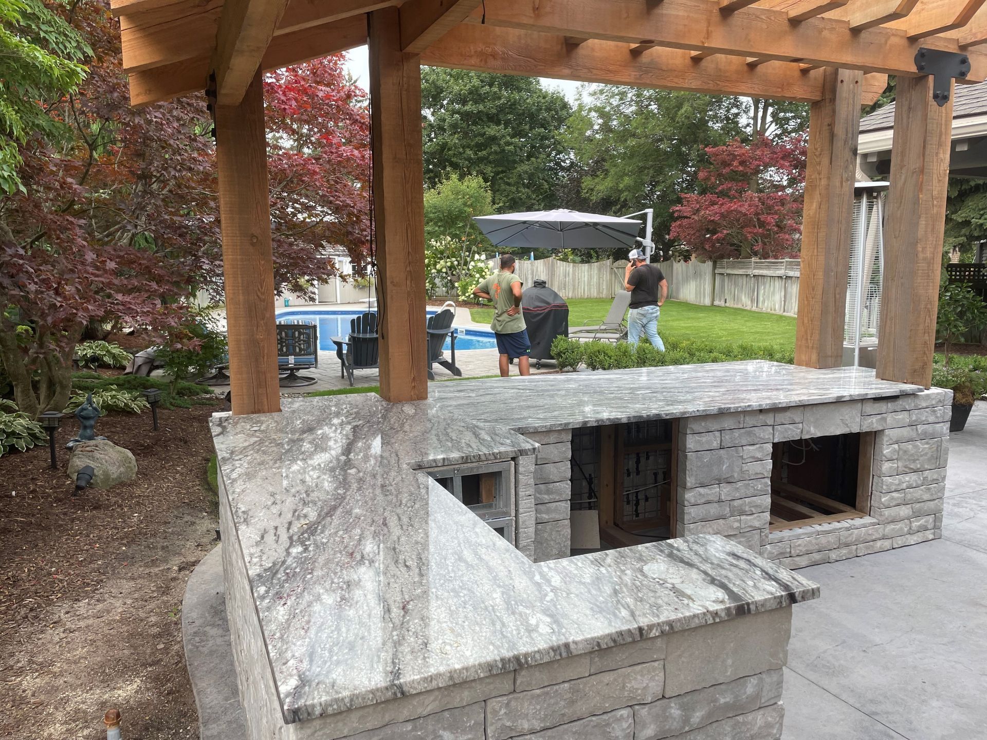 Outdoor kitchen with stone counters and pergola, overlooking a pool and yard. People are present.