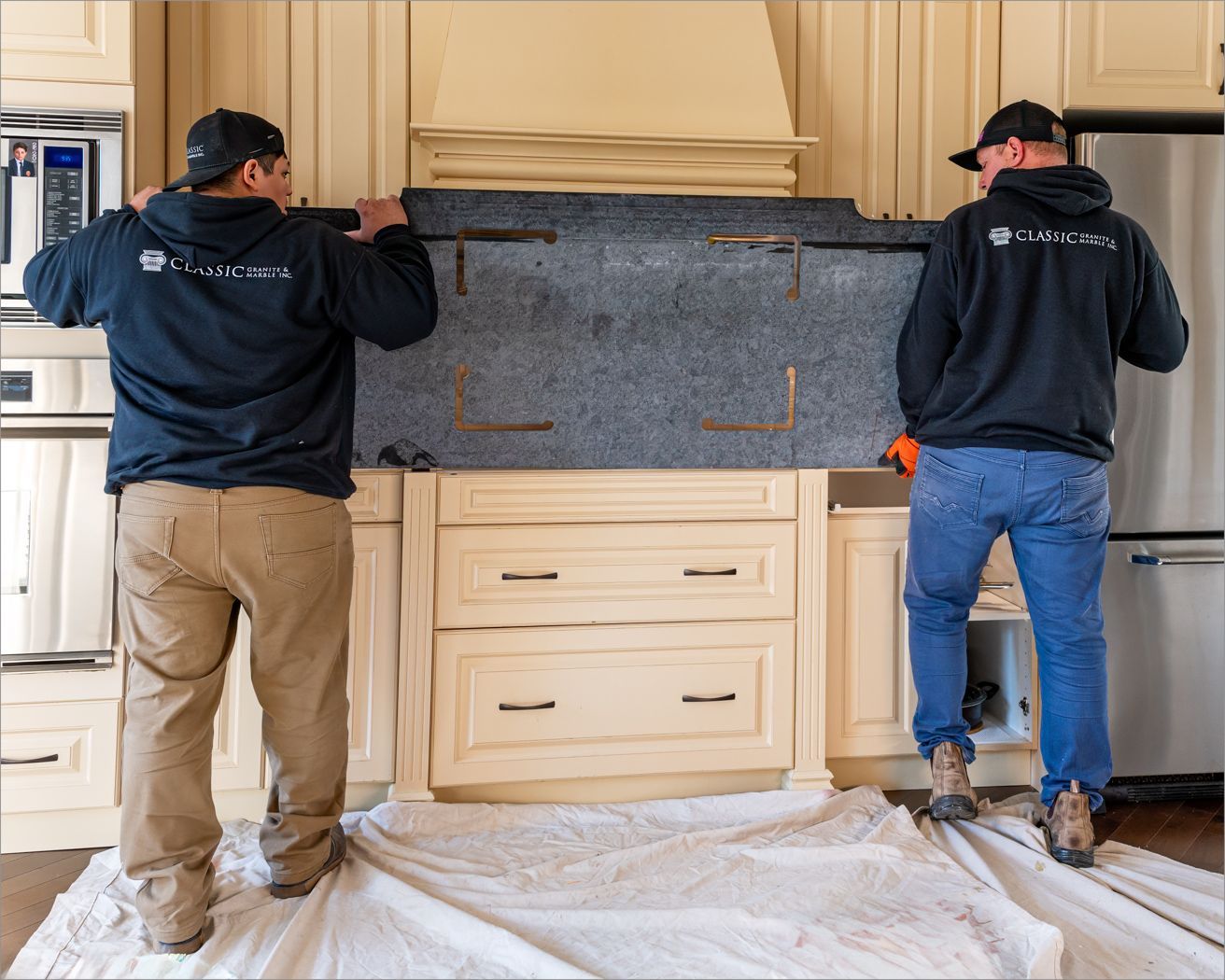 Two men install a stone slab above kitchen cabinets. They wear jackets, hold the slab, and stand on a drop cloth.