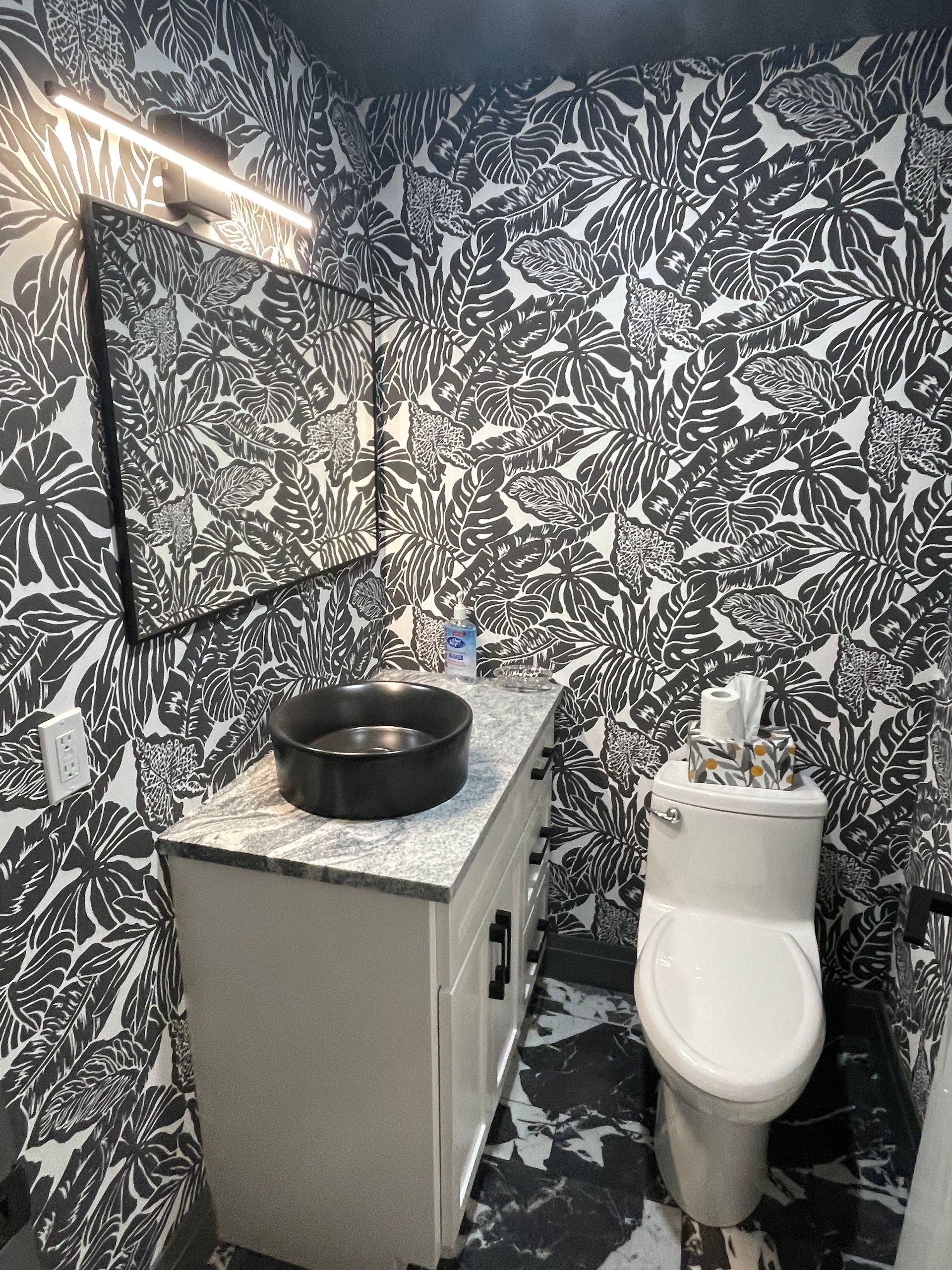 A bathroom with a toilet , sink , mirror and black and white wallpaper.