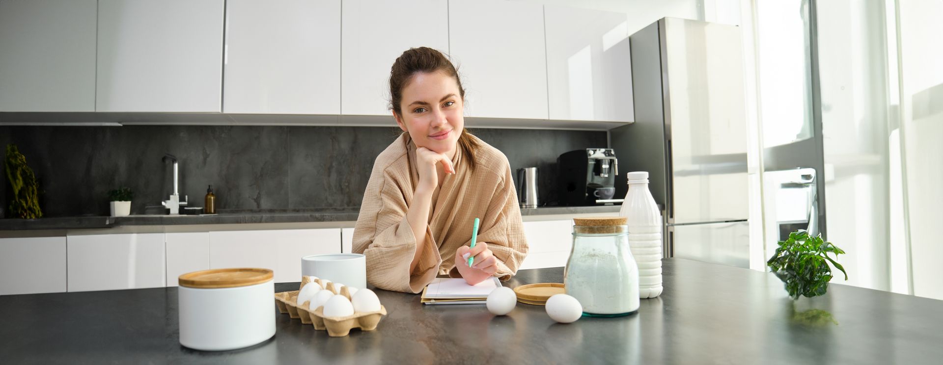 A woman is sitting at a table in a kitchen writing in a notebook.