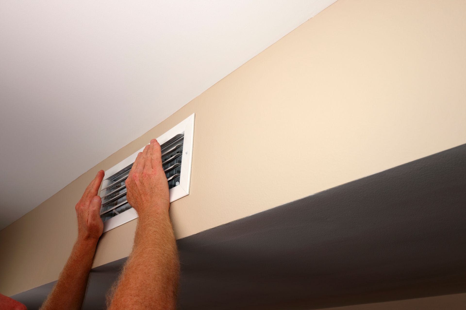 Hands adjusting a white rectangular air vent on a beige wall near a white ceiling.