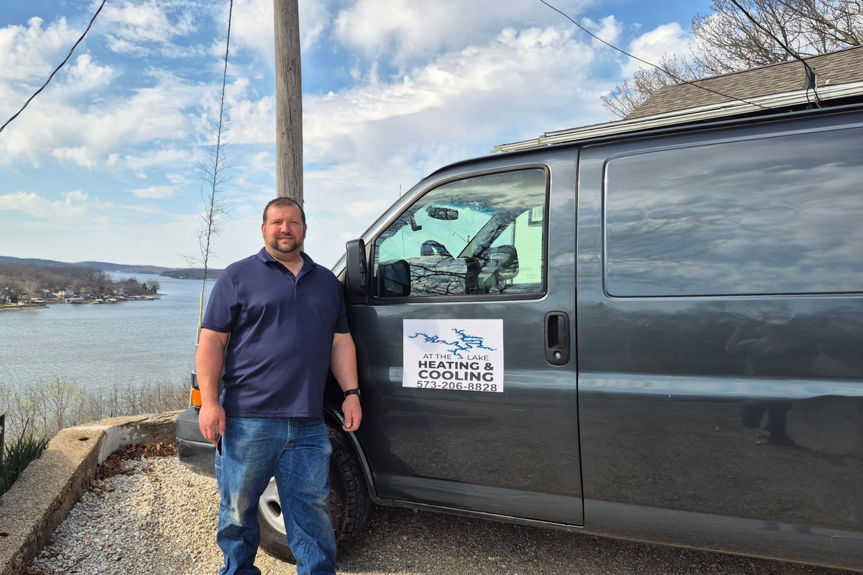 Man standing beside a dark van by a waterfront road on a cloudy day