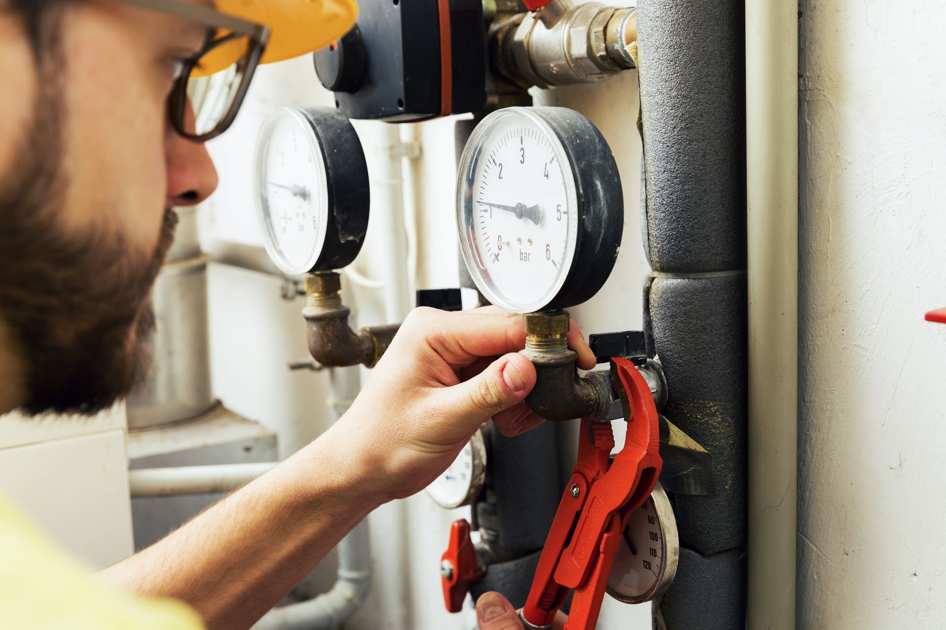 A technician in a yellow hat adjusts gauges on a pipe with red pliers.