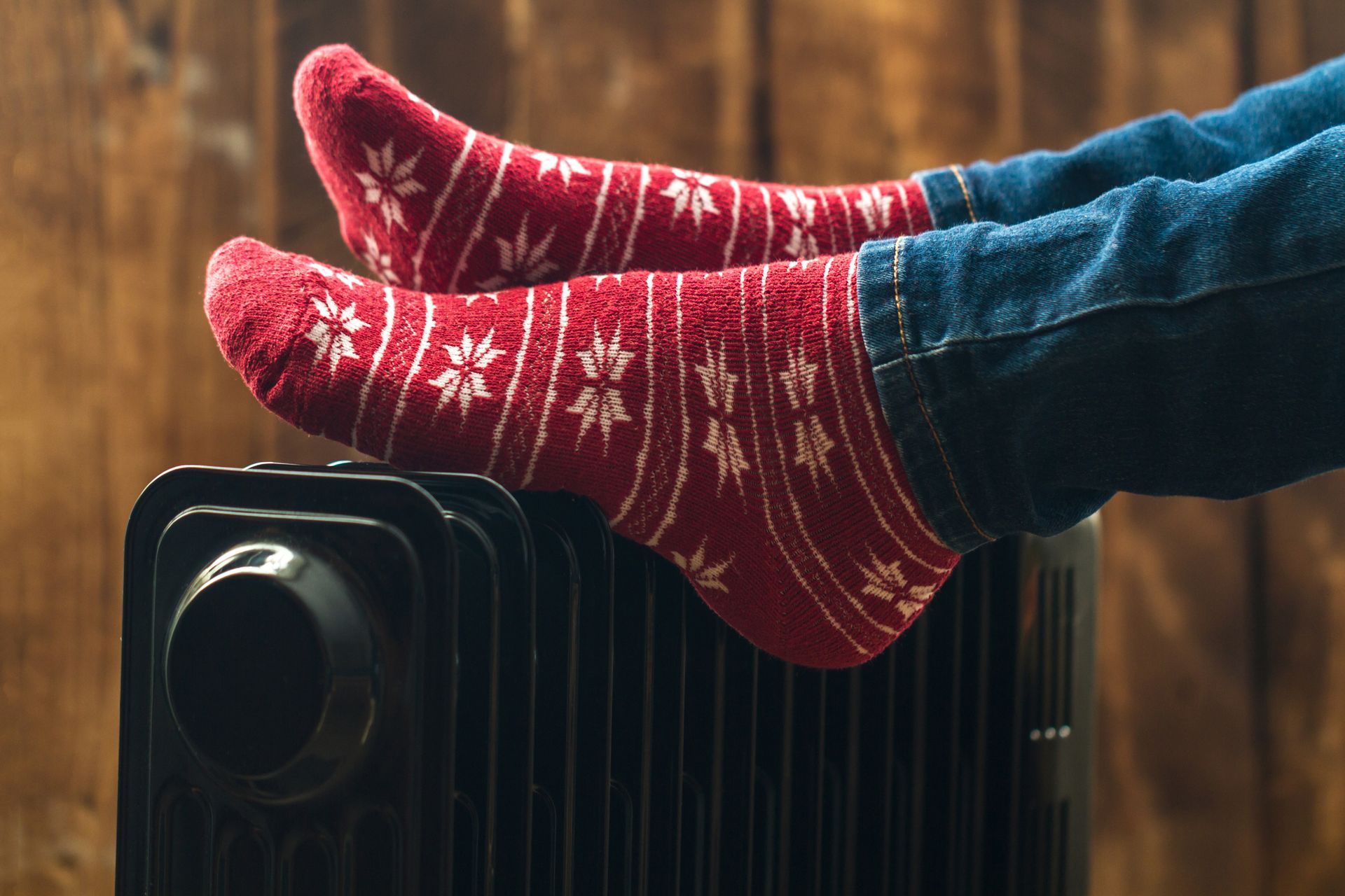 Feet in red snowflake socks resting on a black radiator, wood background.
