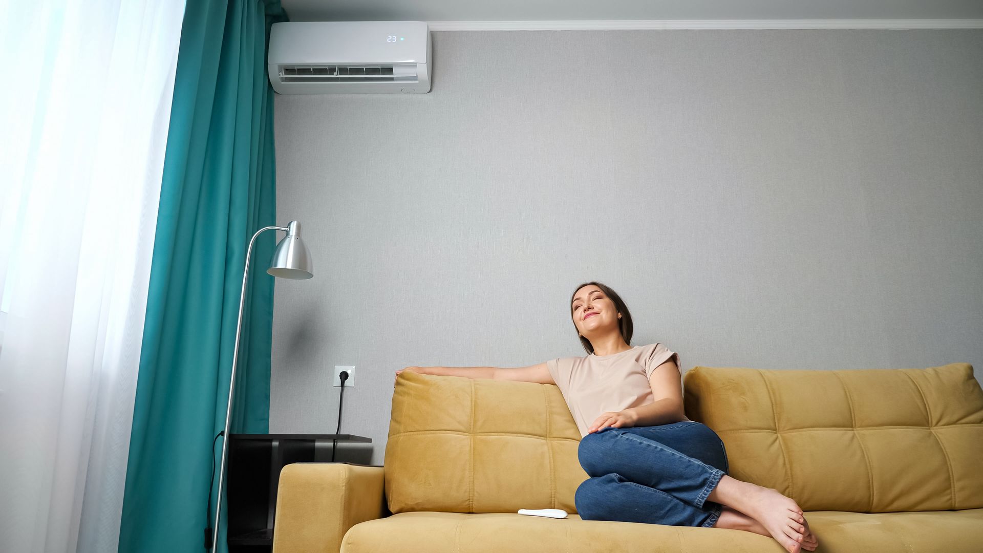 Person reclining on a mustard-yellow sofa in a bright living room, looking upward near a window and air conditioner.