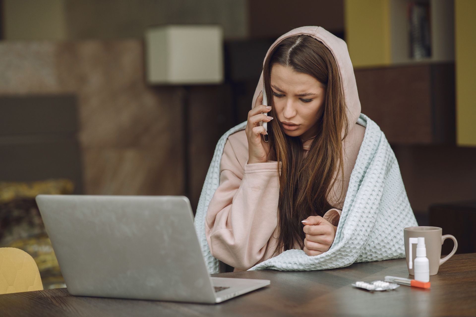 A woman wrapped in a blanket is sitting at a table with a laptop and talking on a cell phone.