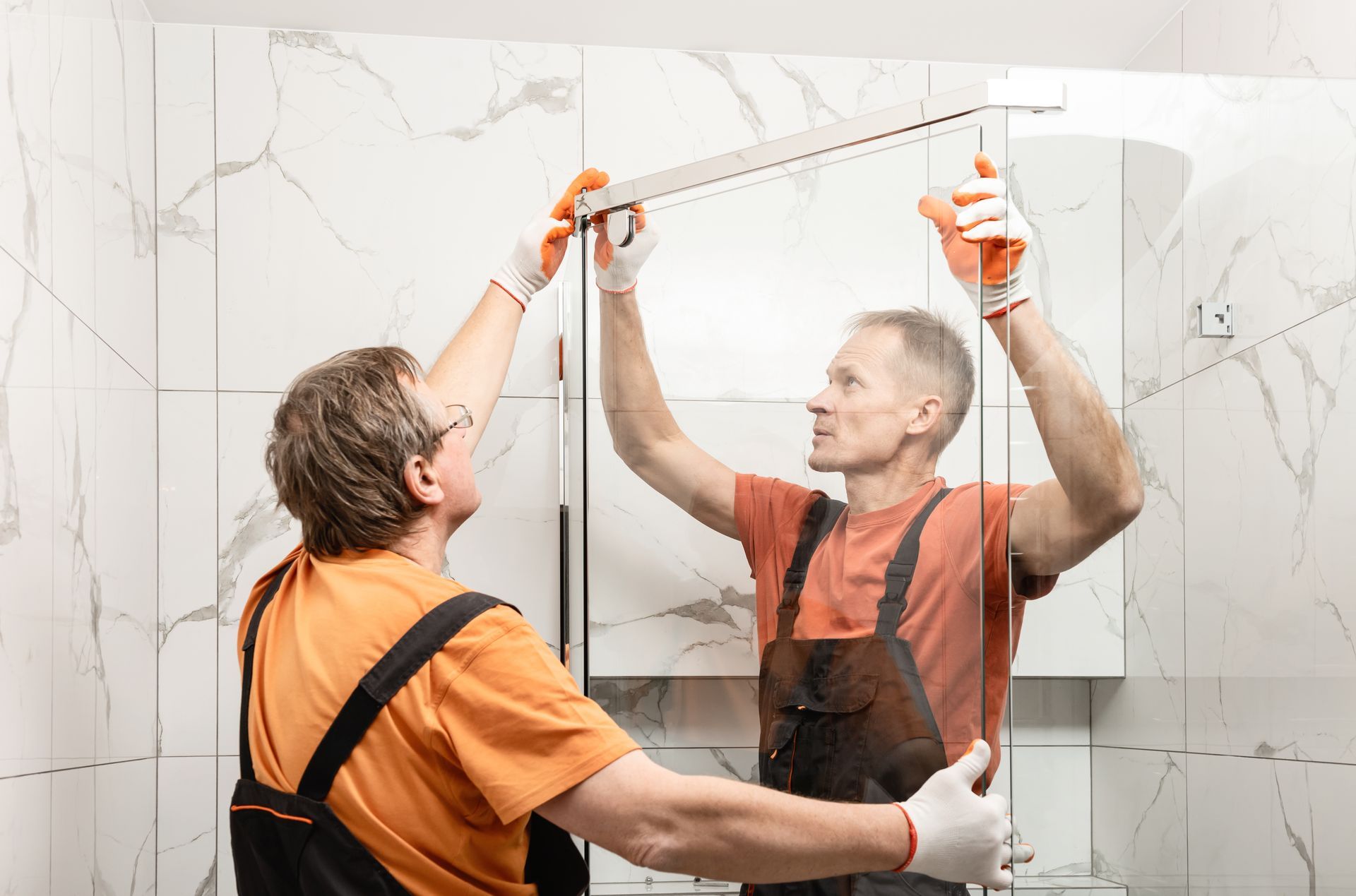 A pair of male workers install a shower door inside a bathroom.