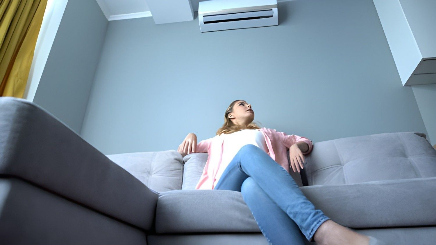 A Woman on A Gray Couch Looking up At an Air Conditioner — Allchin Airconditioning & Refrigeration Electrical in Caloundra, QLD