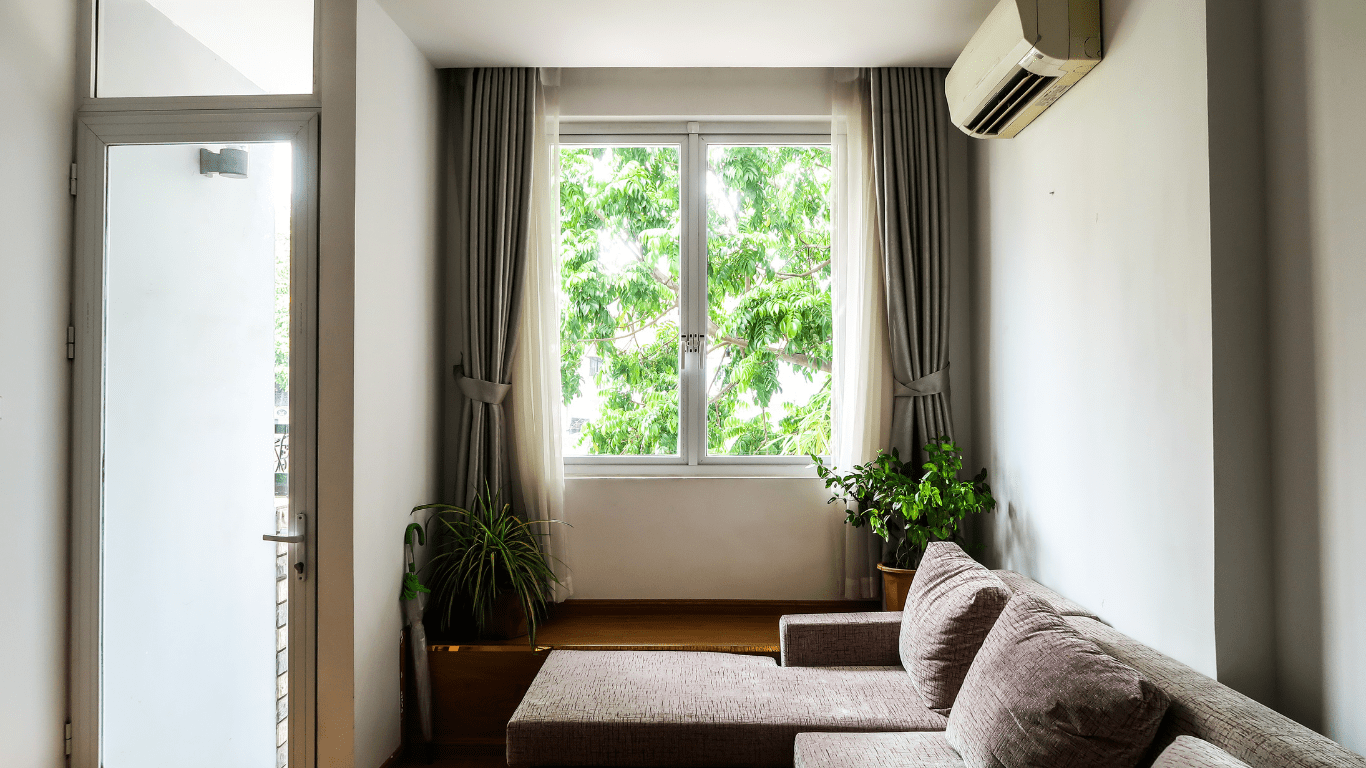 A living room with a window, curtains, sofa, plants, air conditioner — Allchin Airconditioning & Refrigeration Electrical in Caloundra, QLD