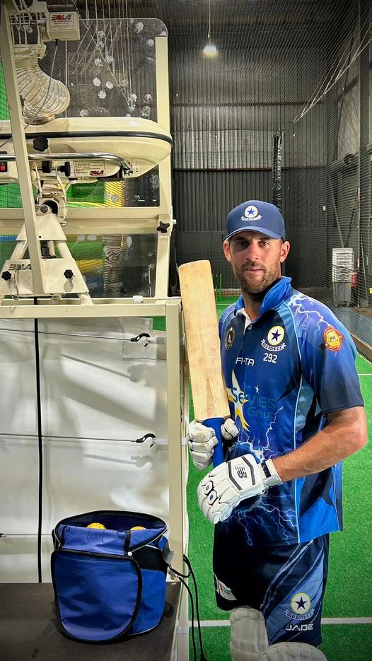 A man in a blue shirt is holding a cricket bat in a cricket stadium.
