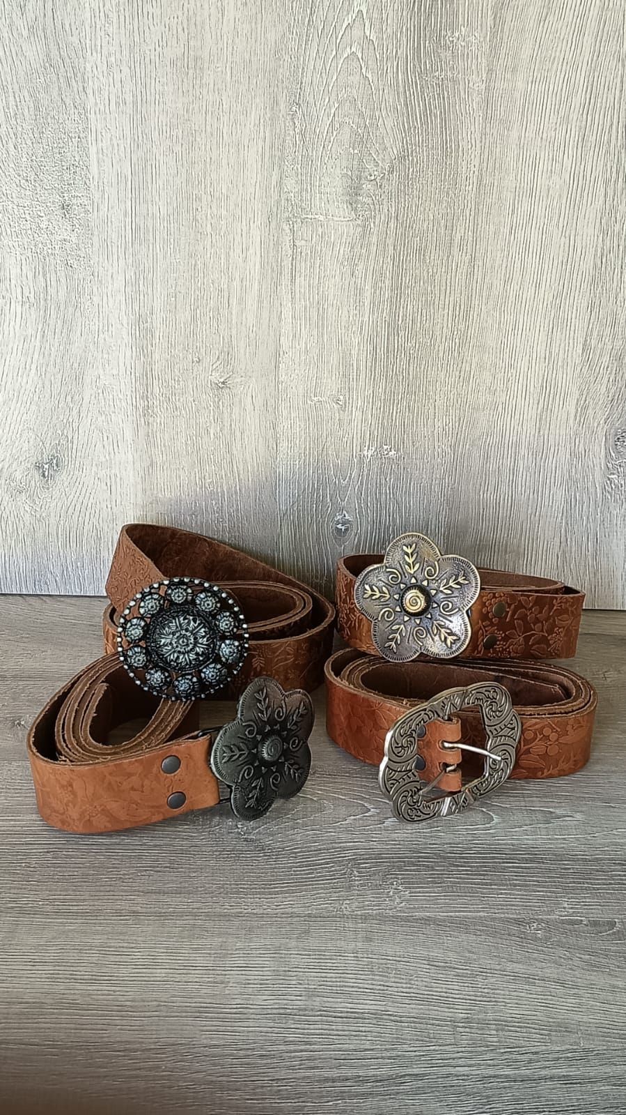 Cinco cinturones de cuero marrón con hebillas plateadas decorativas sobre una superficie de madera.