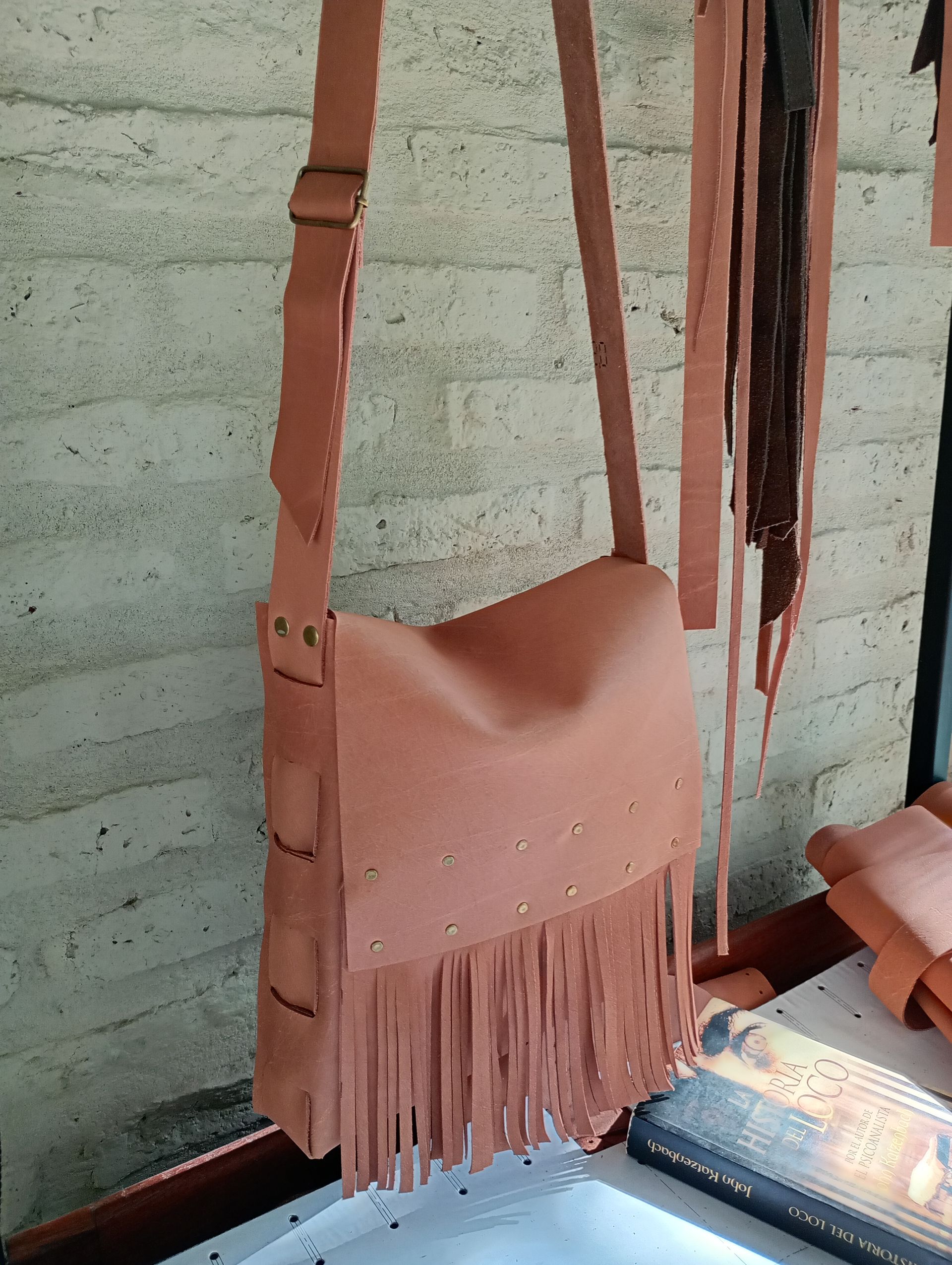 Un bolso de hombro de cuero marrón claro con flecos, colgado contra una pared de ladrillos.