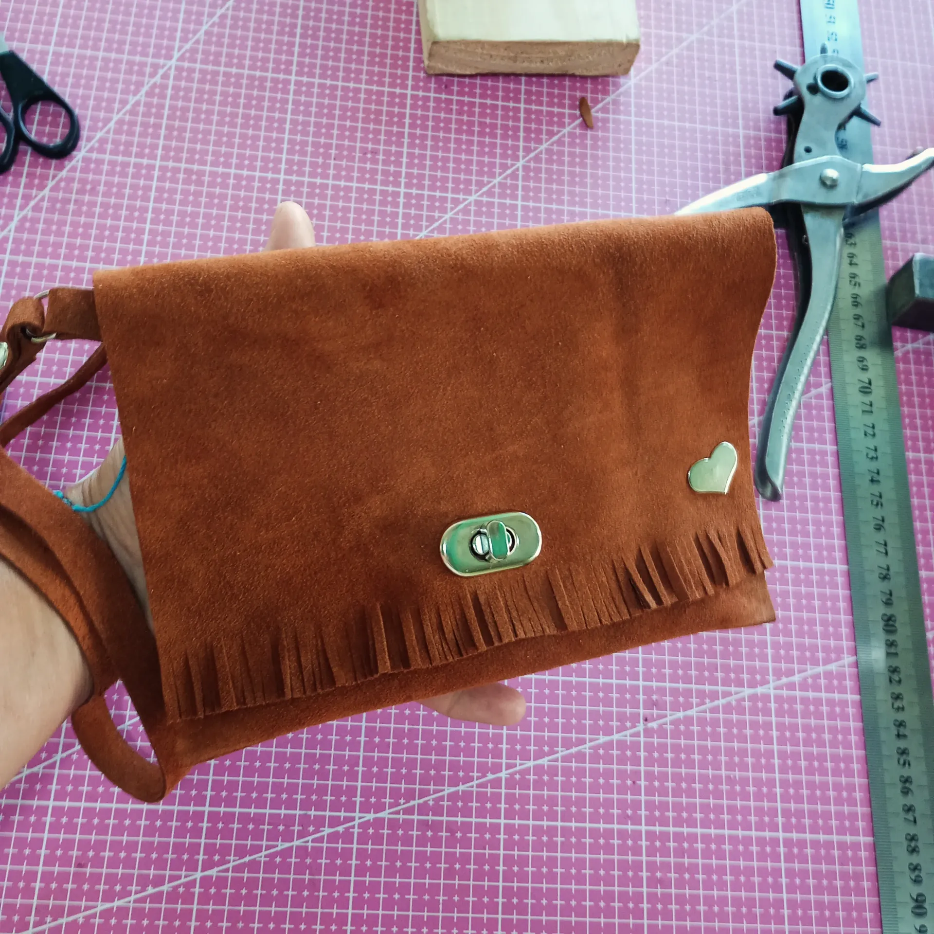 Bolso de ante marrón con flecos, sobre una esterilla de corte rosa. Está rodeado de herramientas.