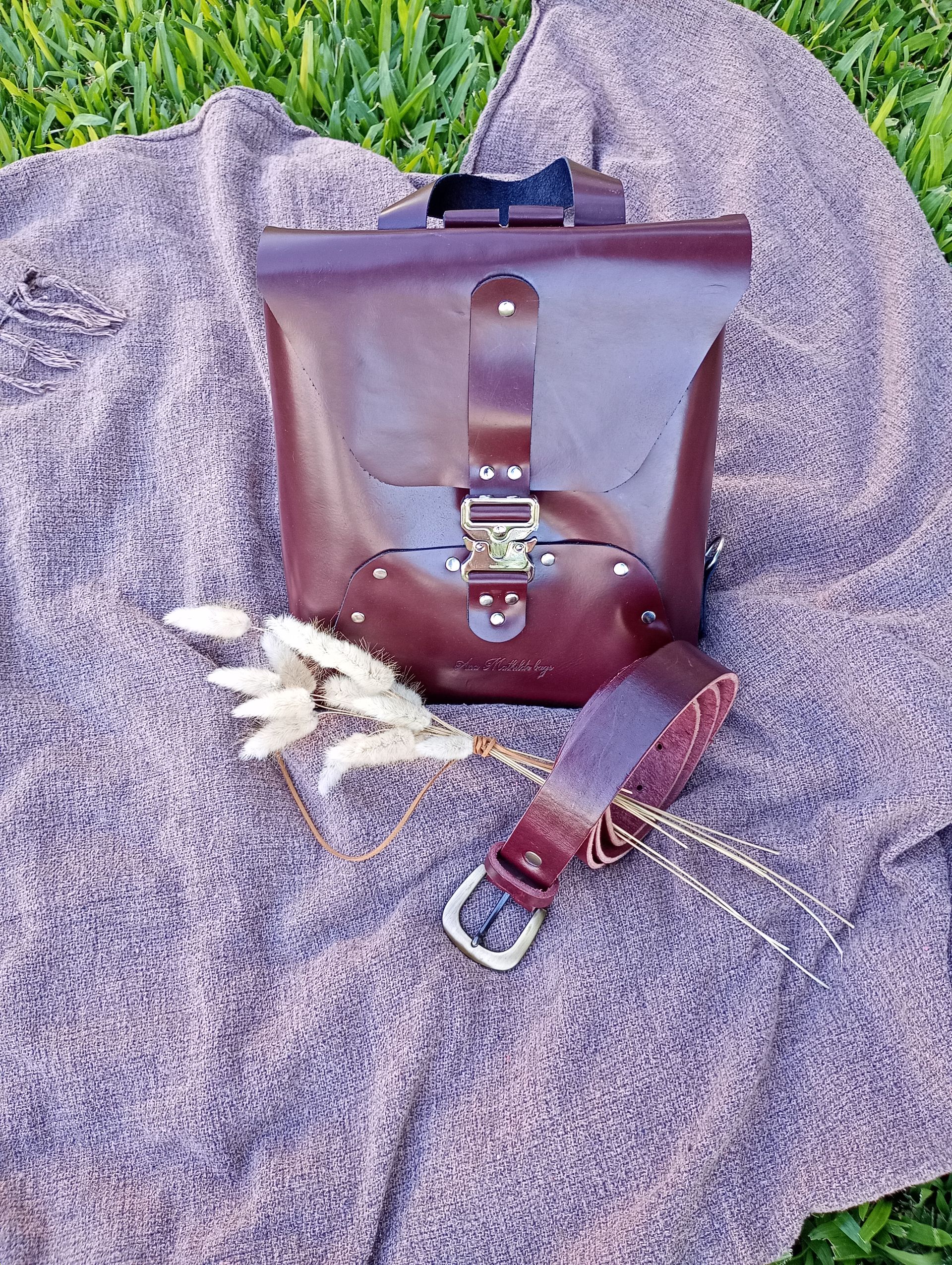 Un bolso y cinturón de cuero marrón con herrajes plateados sobre una manta gris y césped.