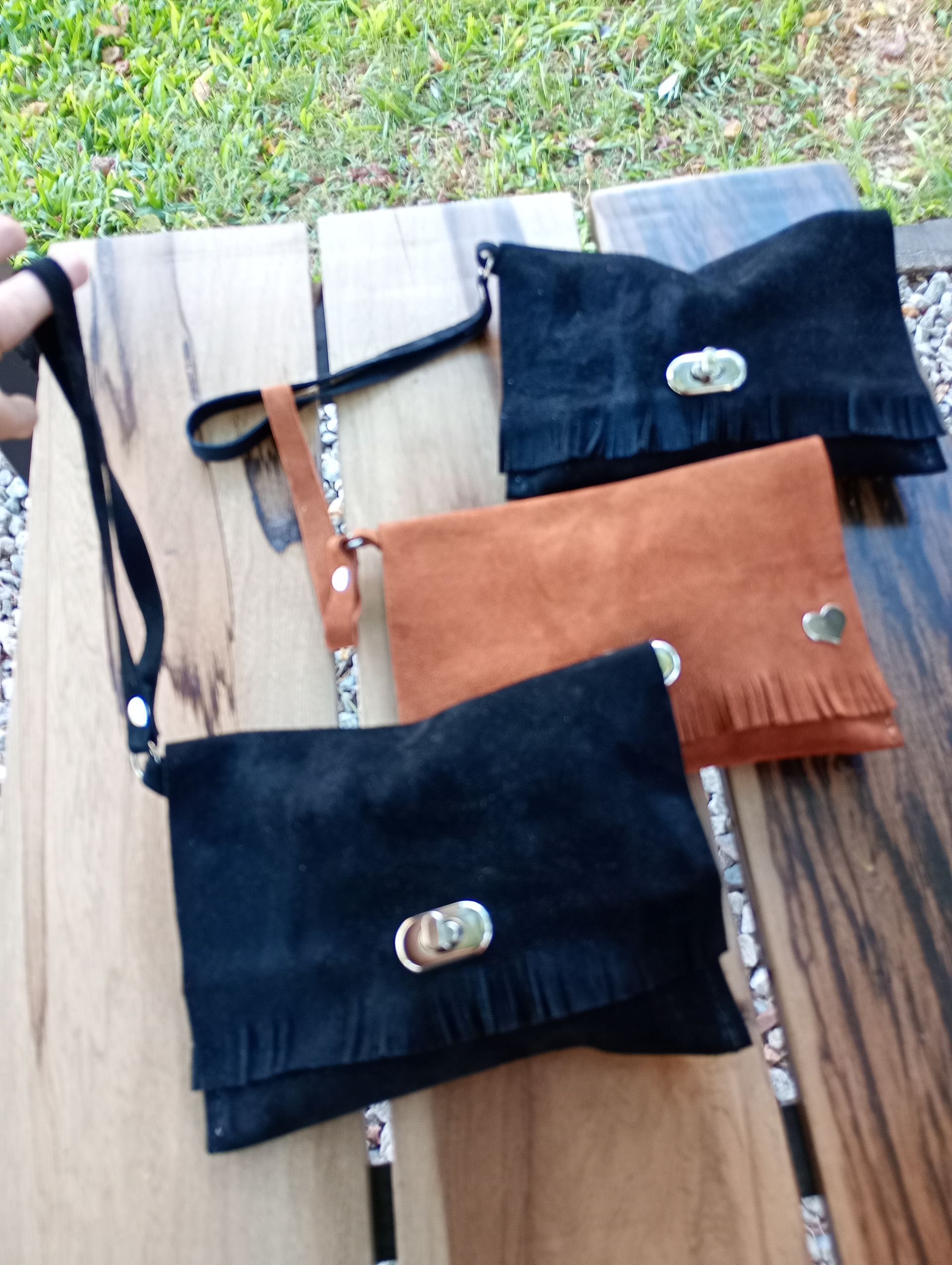 Tres bolsos de mano de ante en color negro y marrón, con flecos y correas de muñeca, expuestos sobre madera.