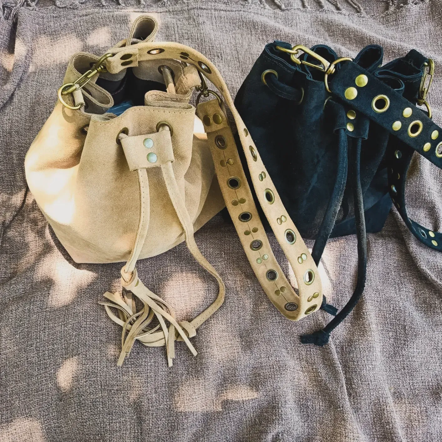 Dos bolsos tipo cubo de ante, uno beige y otro azul marino, con cierres de cordón y correas con tachuelas.