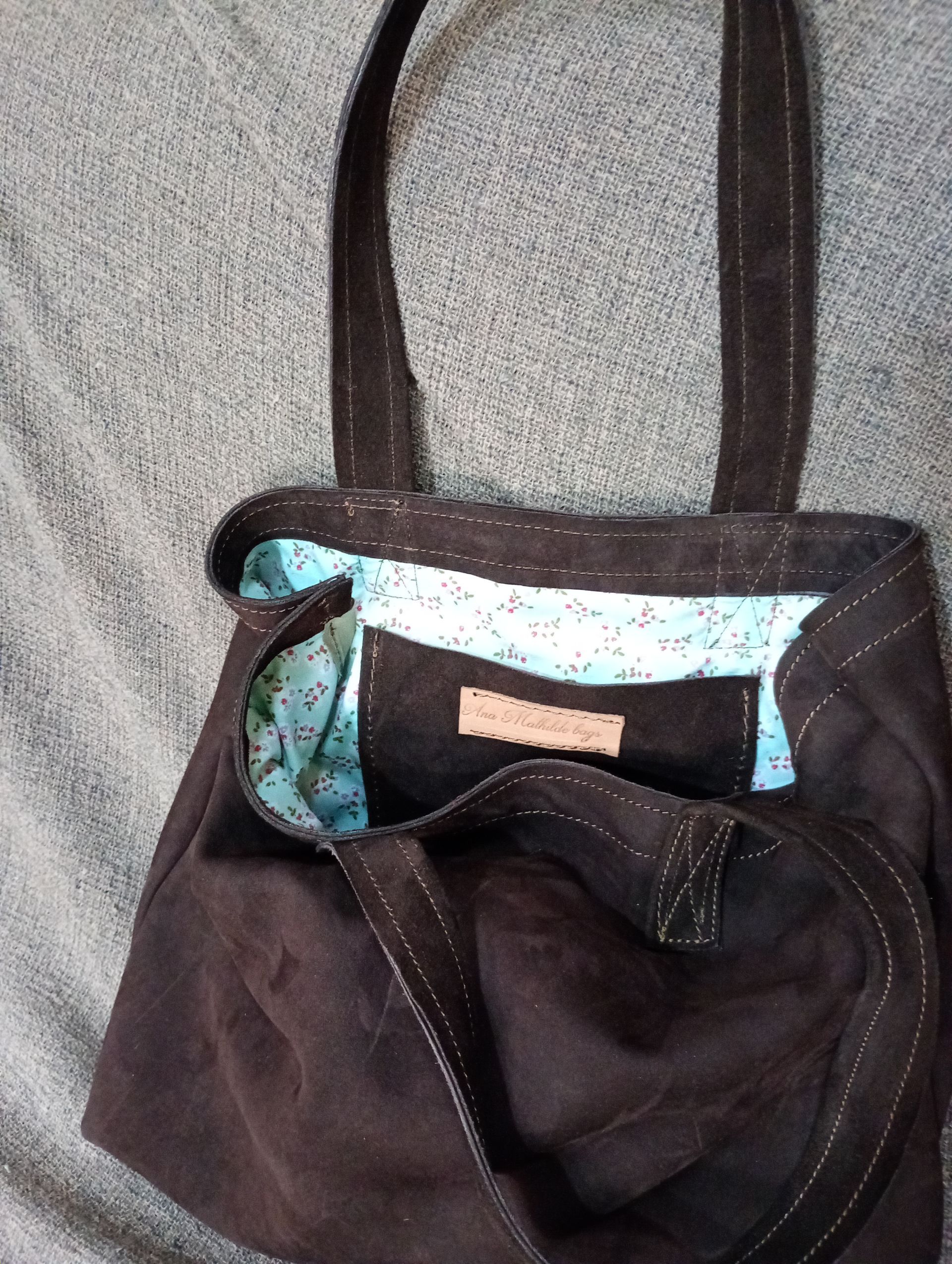Bolso tote de color marrón oscuro con forro estampado en azul, sobre una superficie gris.