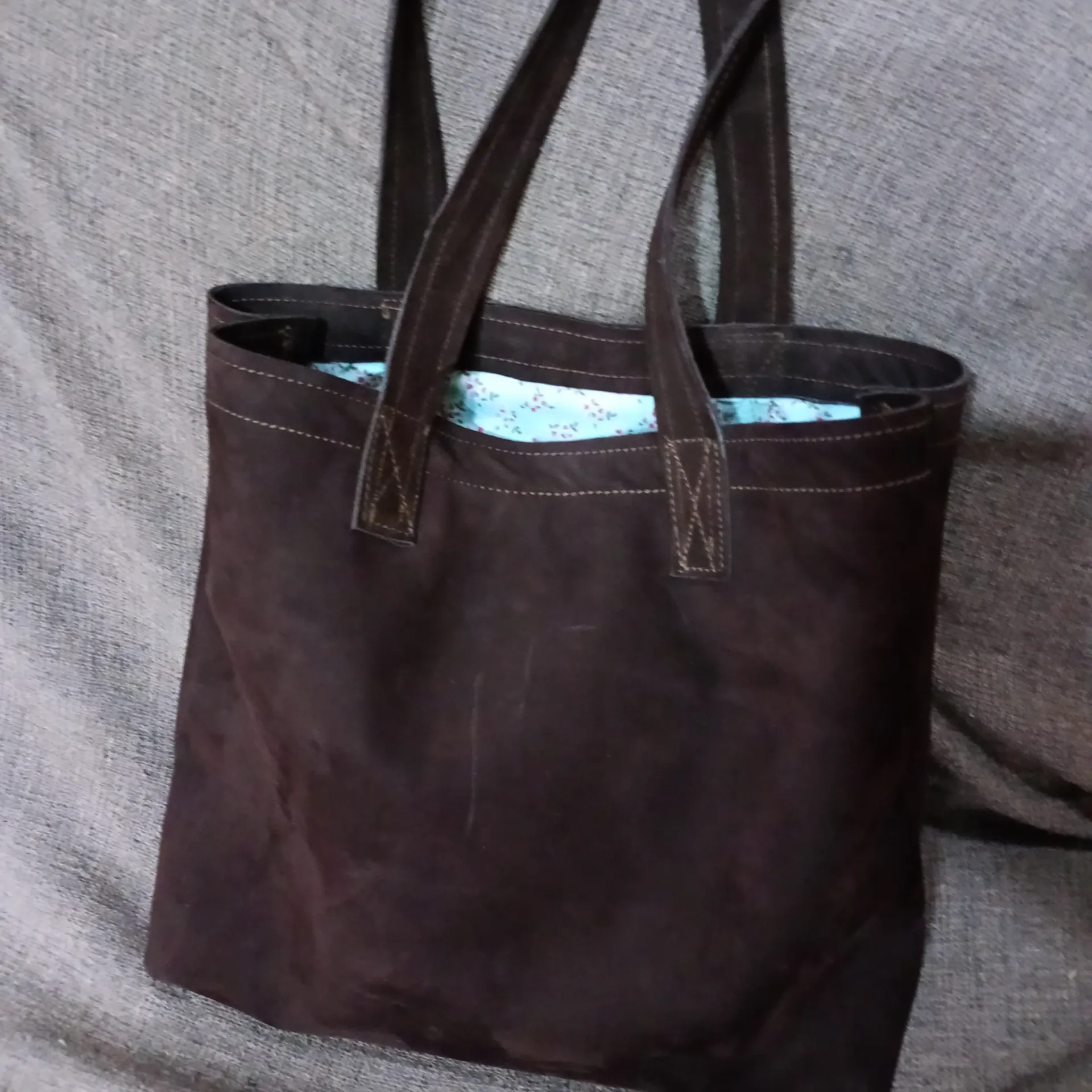 Bolso tote de color marrón oscuro con forro estampado en azul, sobre una superficie gris.
