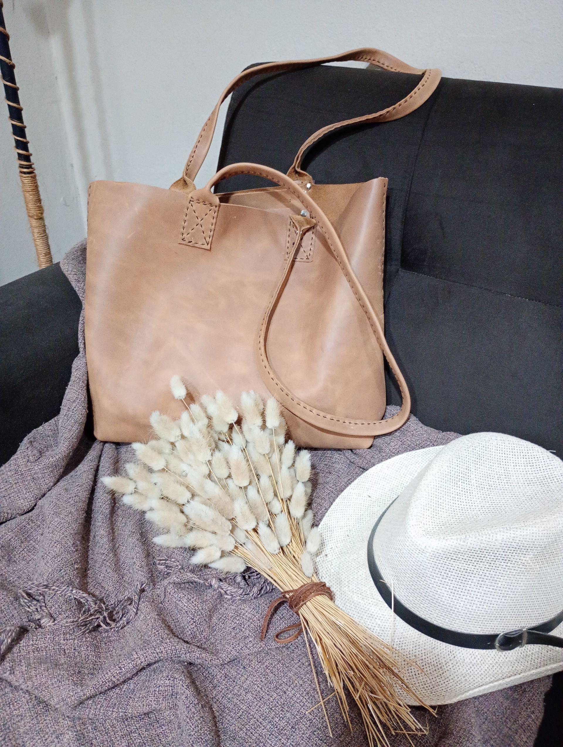 Bolso tote de piel beige con ramo y sombrero sobre manta morada.
