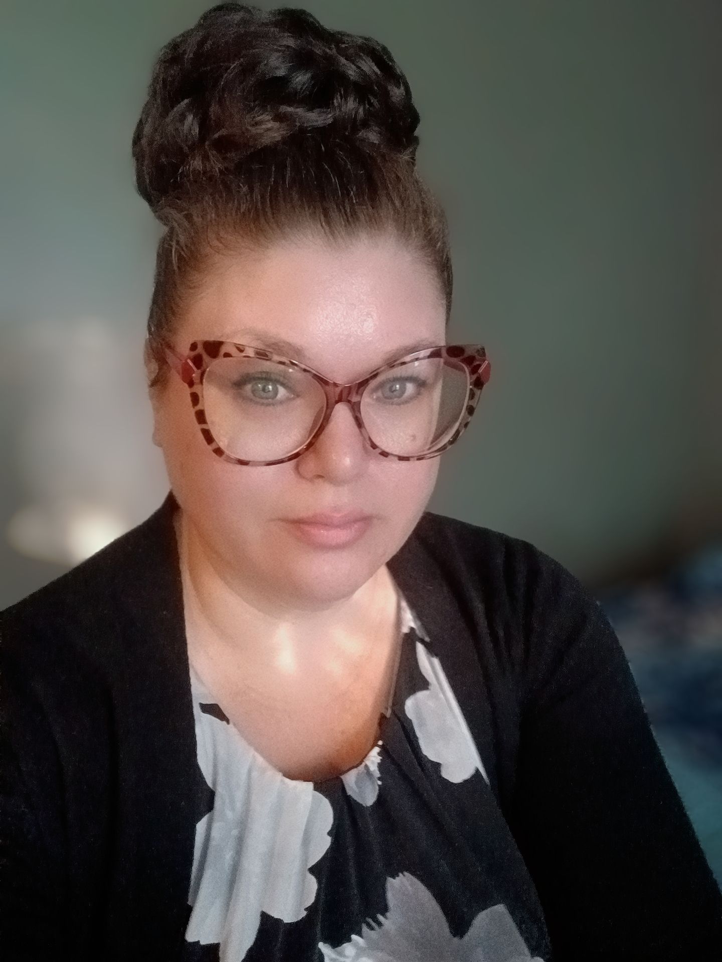 Woman with dark hair in a bun, wearing large, patterned glasses and a black cardigan over a floral top.
