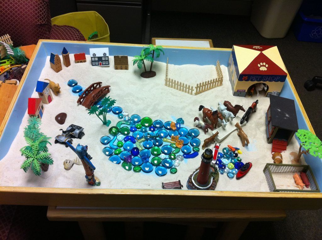 A sand tray scene with miniature houses, animals, trees, and water made of blue glass stones.