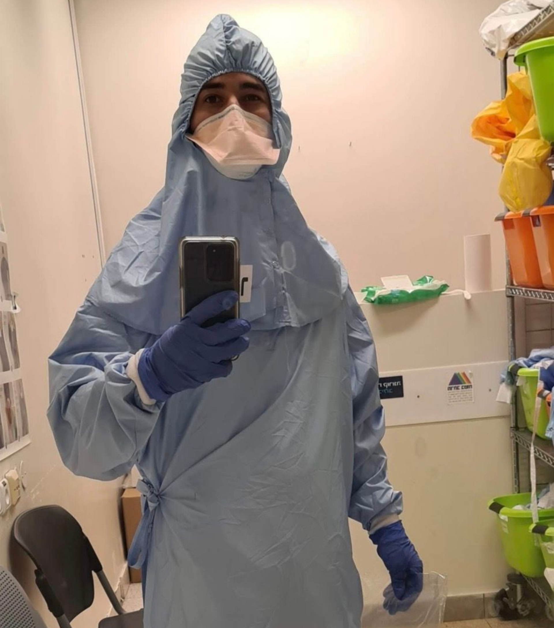 A person in a blue medical gown, hood, and face mask taking a selfie in a storage room with shelves of medical supplies.