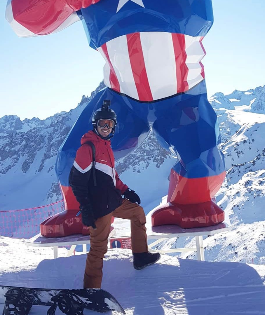 A person in a red and black ski jacket stands smiling on a snowy mountain beneath a large Captain America statue.