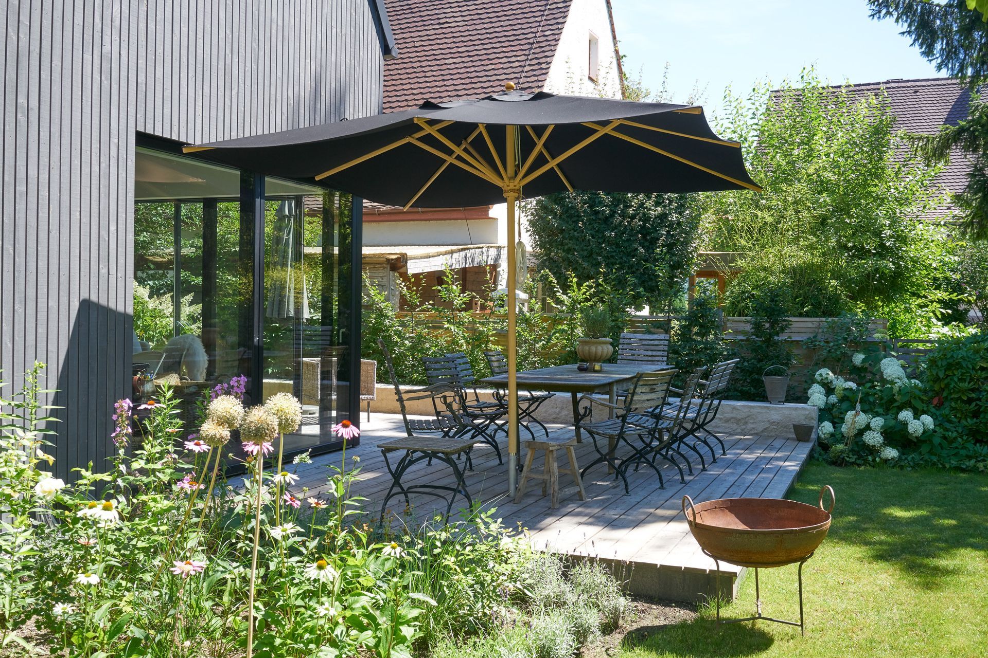 Patio with black umbrella, table, chairs, and garden with flowers and foliage.