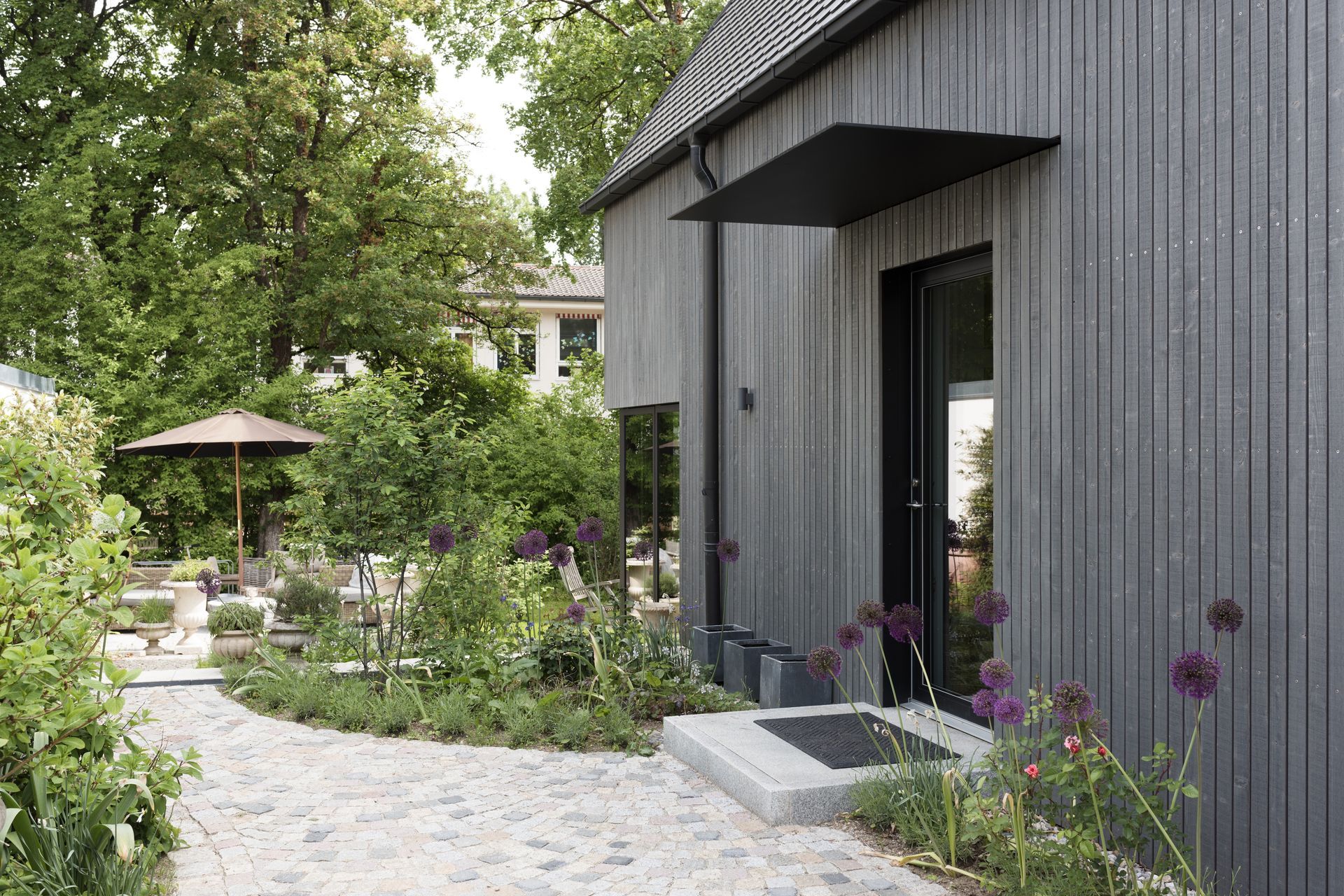 A gray-sided building with a black awning and door, set in a lush garden with gravel path and outdoor seating.