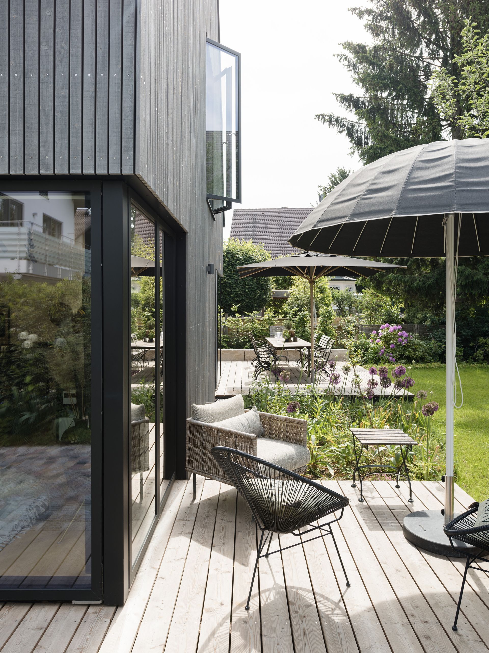 Black-sided modern home with outdoor seating, wooden deck, umbrella, and a lush garden.