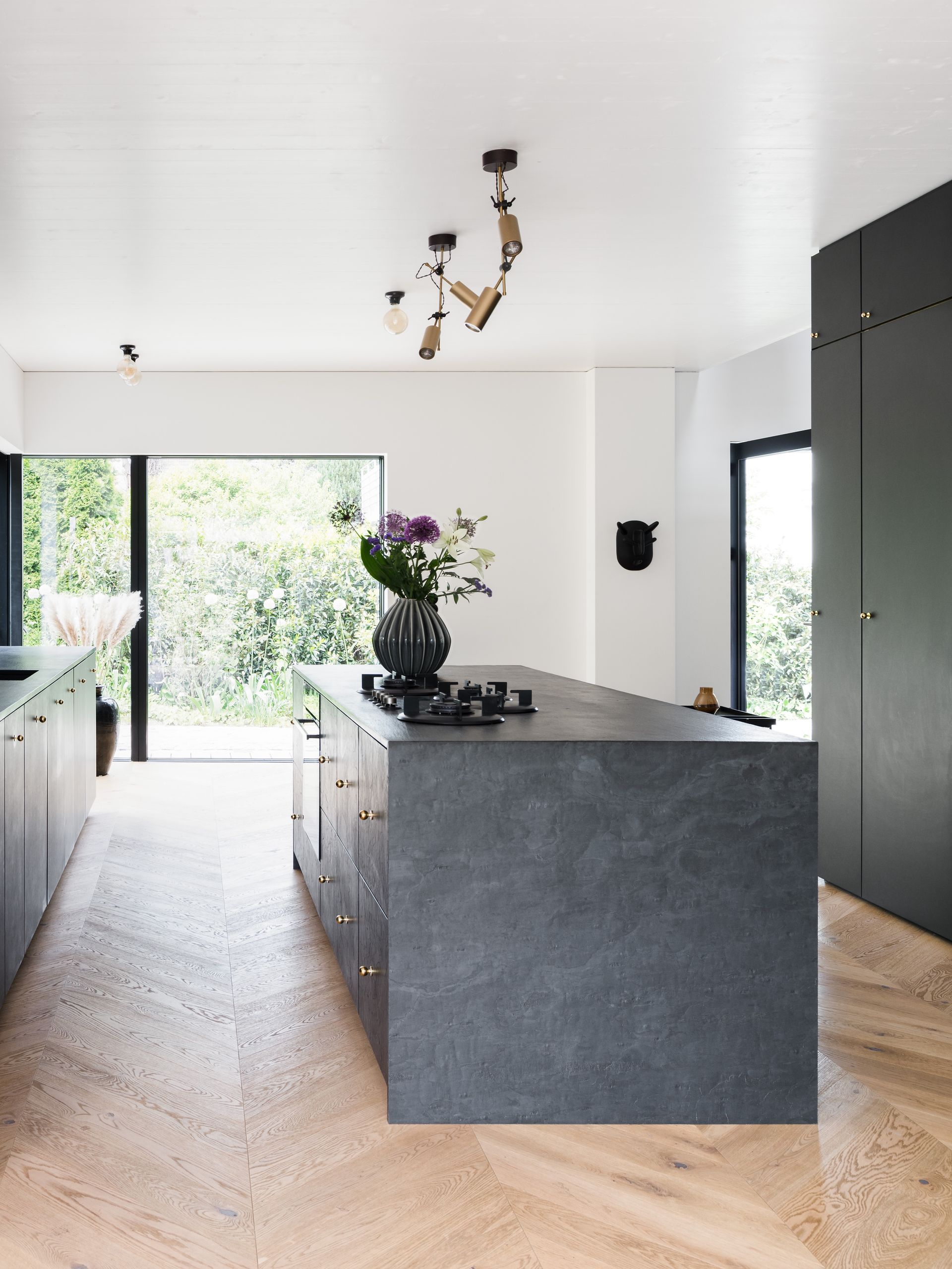 Modern kitchen with a gray island, light wood floor, and large window overlooking greenery.