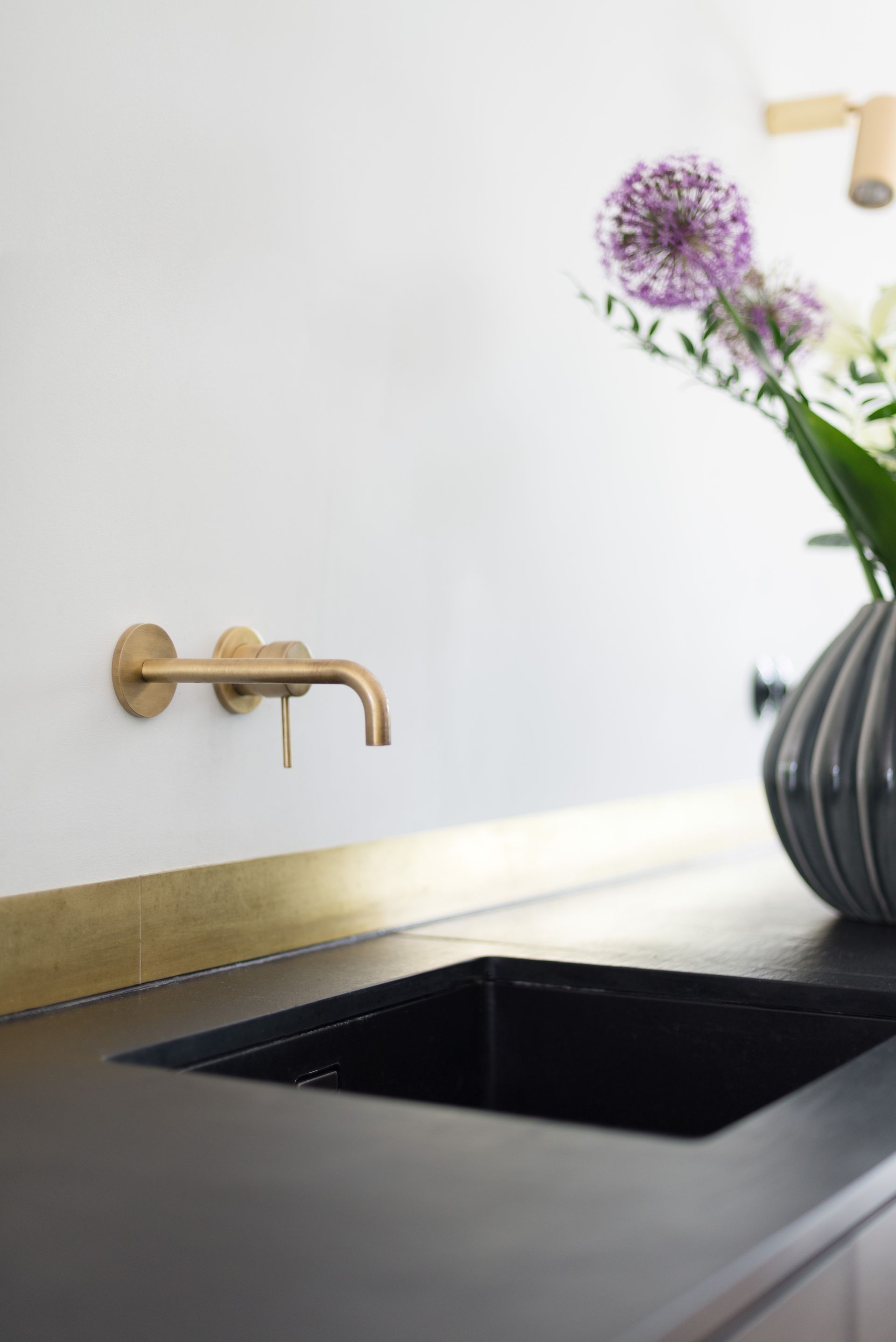 Gold wall-mounted faucet over a black sink with a vase of purple flowers and a gold countertop accent against a white wall.