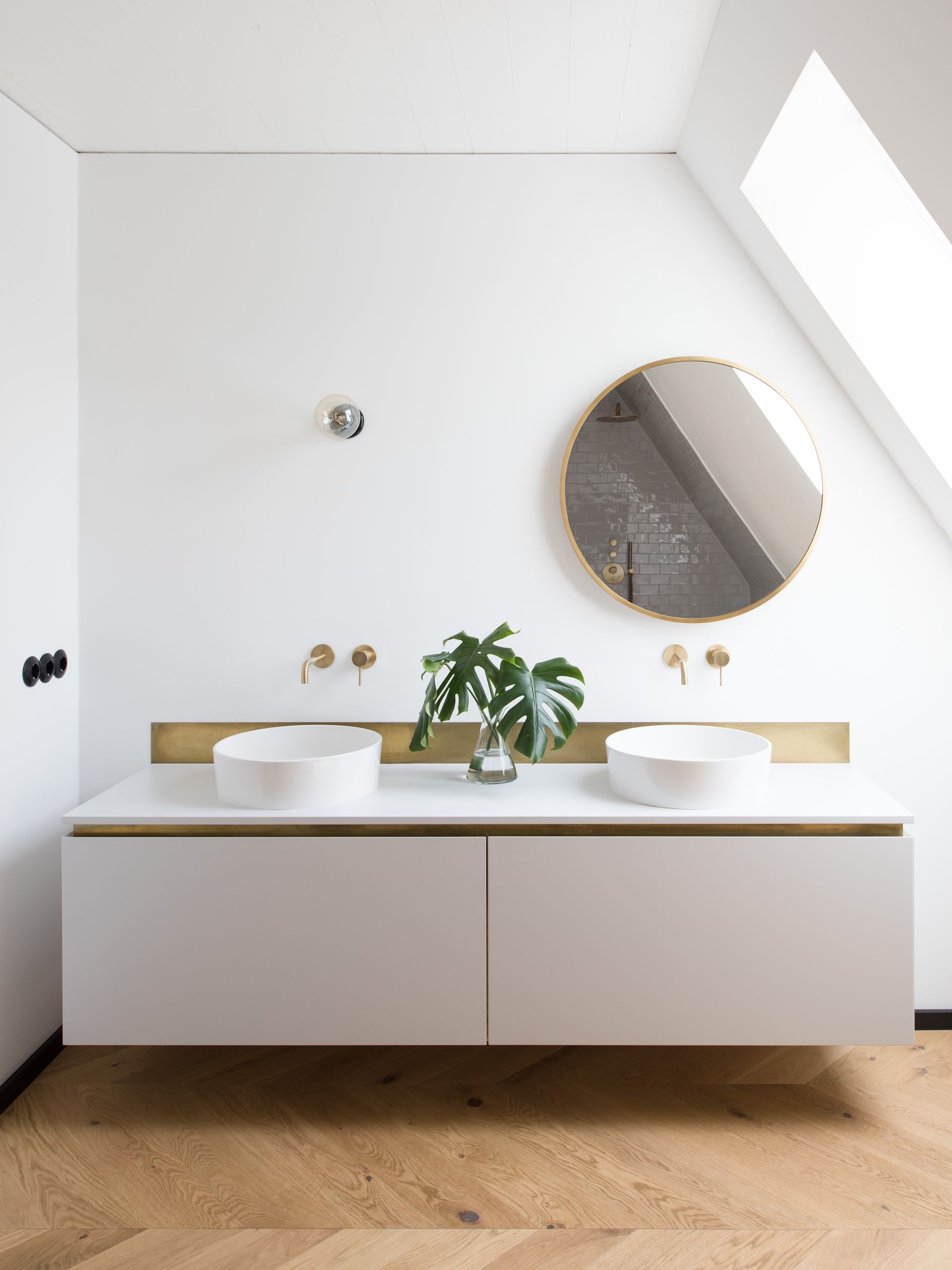 Modern white bathroom with double sink, gold accents, round mirror, and wooden floor.