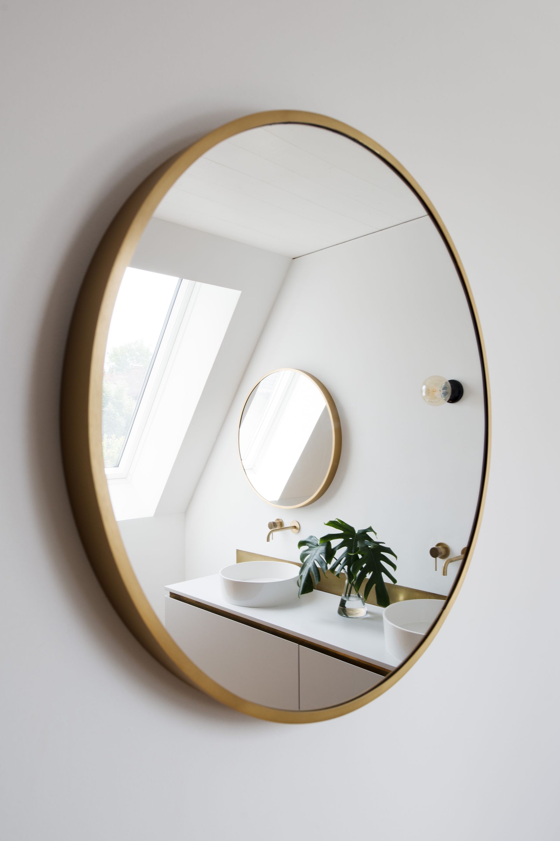Round gold-framed mirror on white wall, reflecting a bathroom with sink, skylight, and smaller mirror.