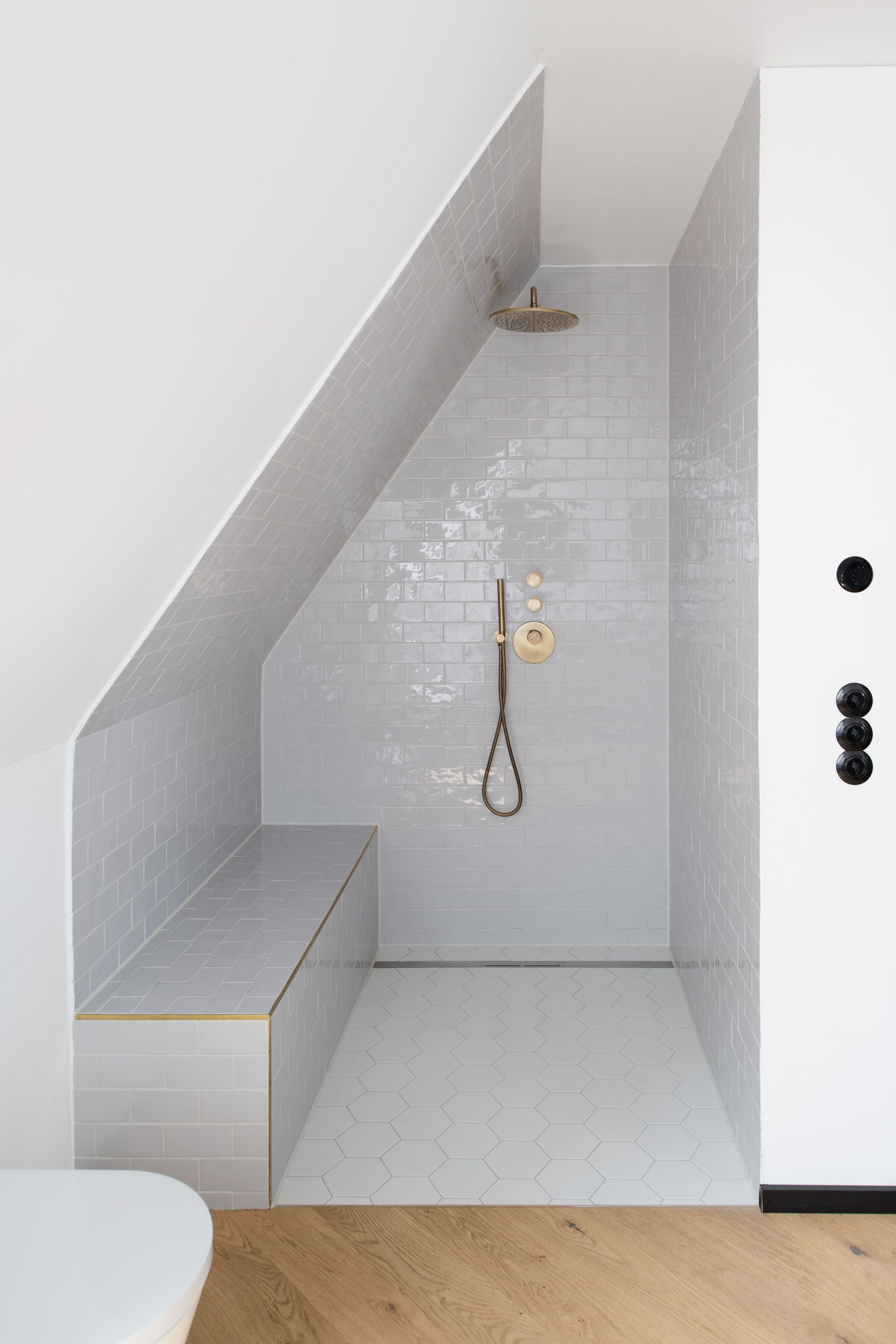 Shower in a small bathroom with angled ceiling. Light gray tiles and a built-in bench.