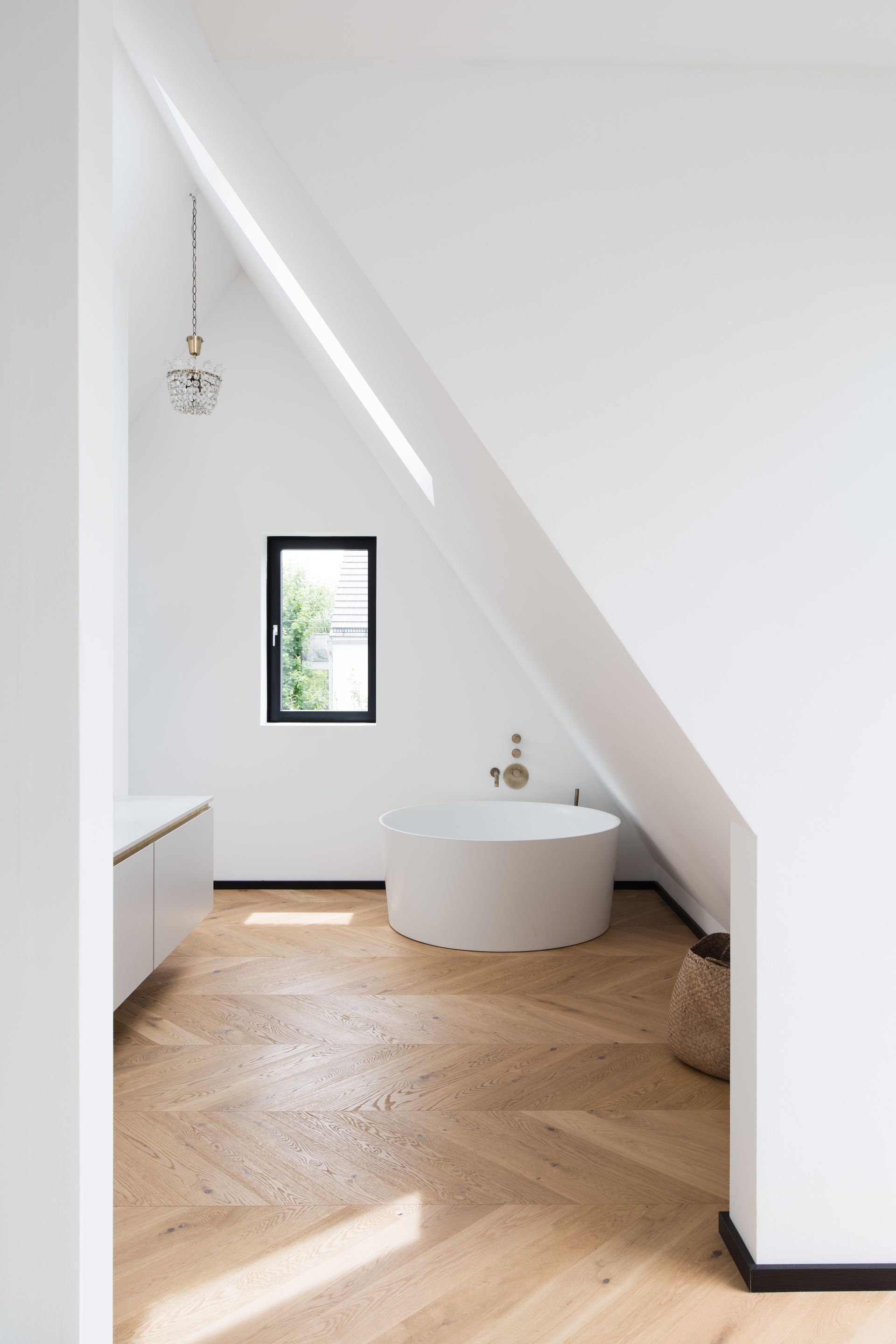 White bathroom with slanted ceiling, small window, circular tub, wood floor.
