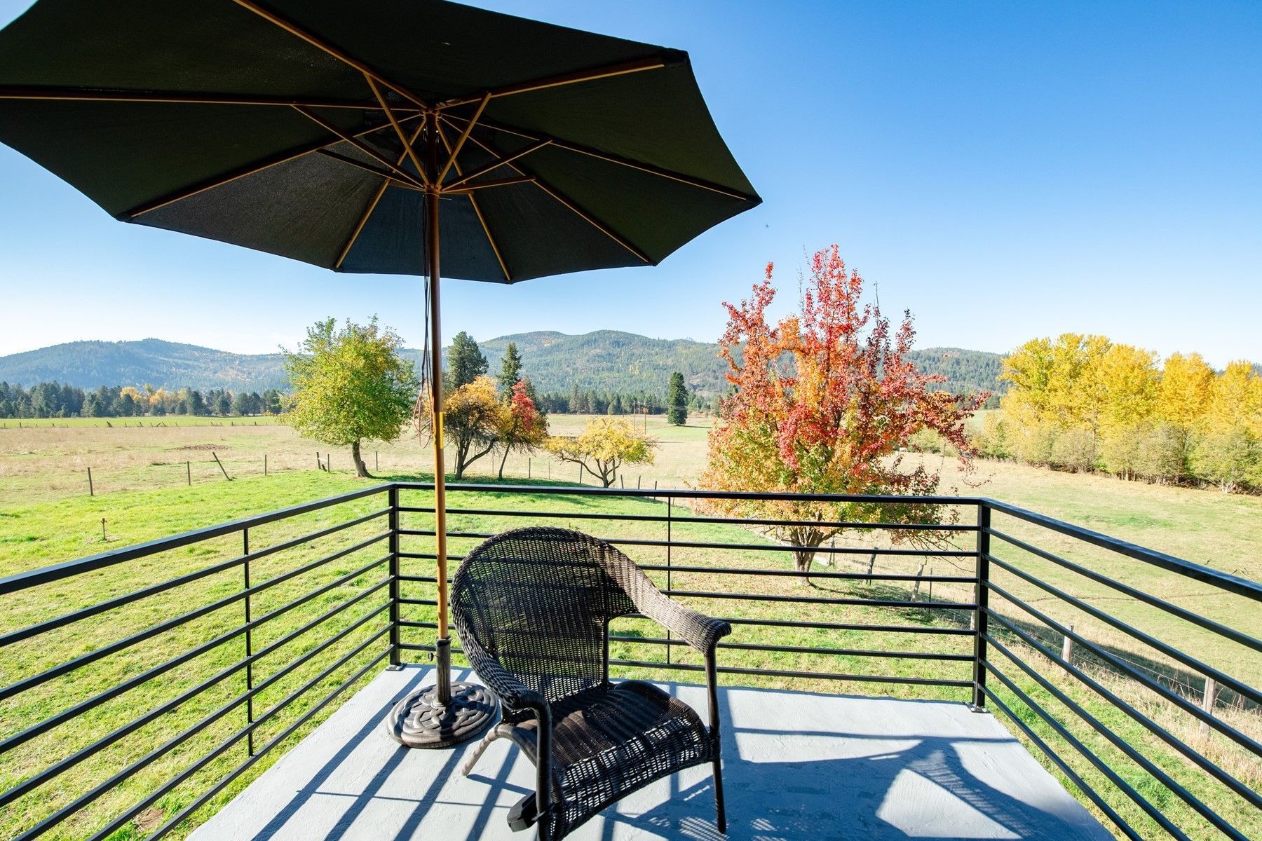 Balcony with black umbrella, chair, overlooking green field and colorful trees. Sunny day.