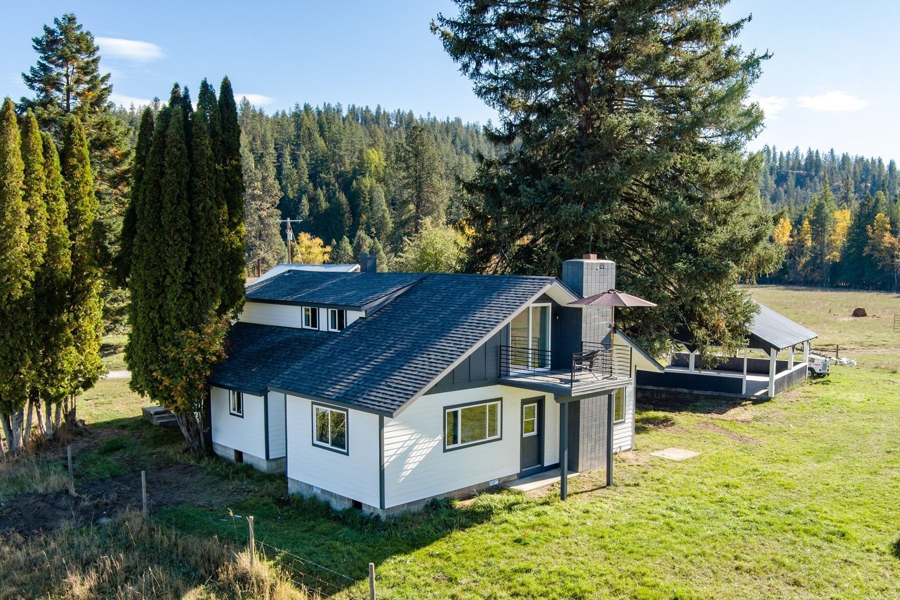 White house with black roof, set in a field with trees and a forest in the background.