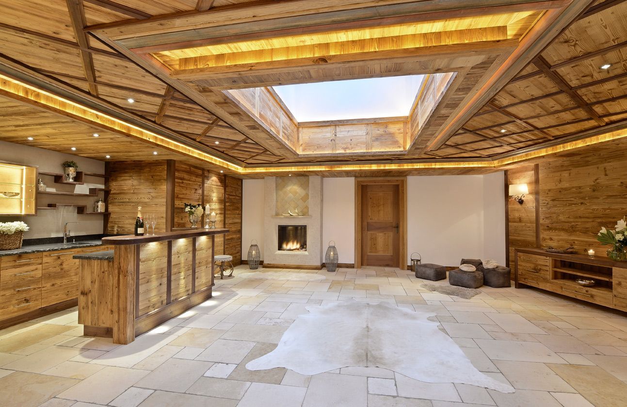 Cozy wooden-paneled room with bar, fireplace, and skylight. Light-colored stone floor and cowhide rug.