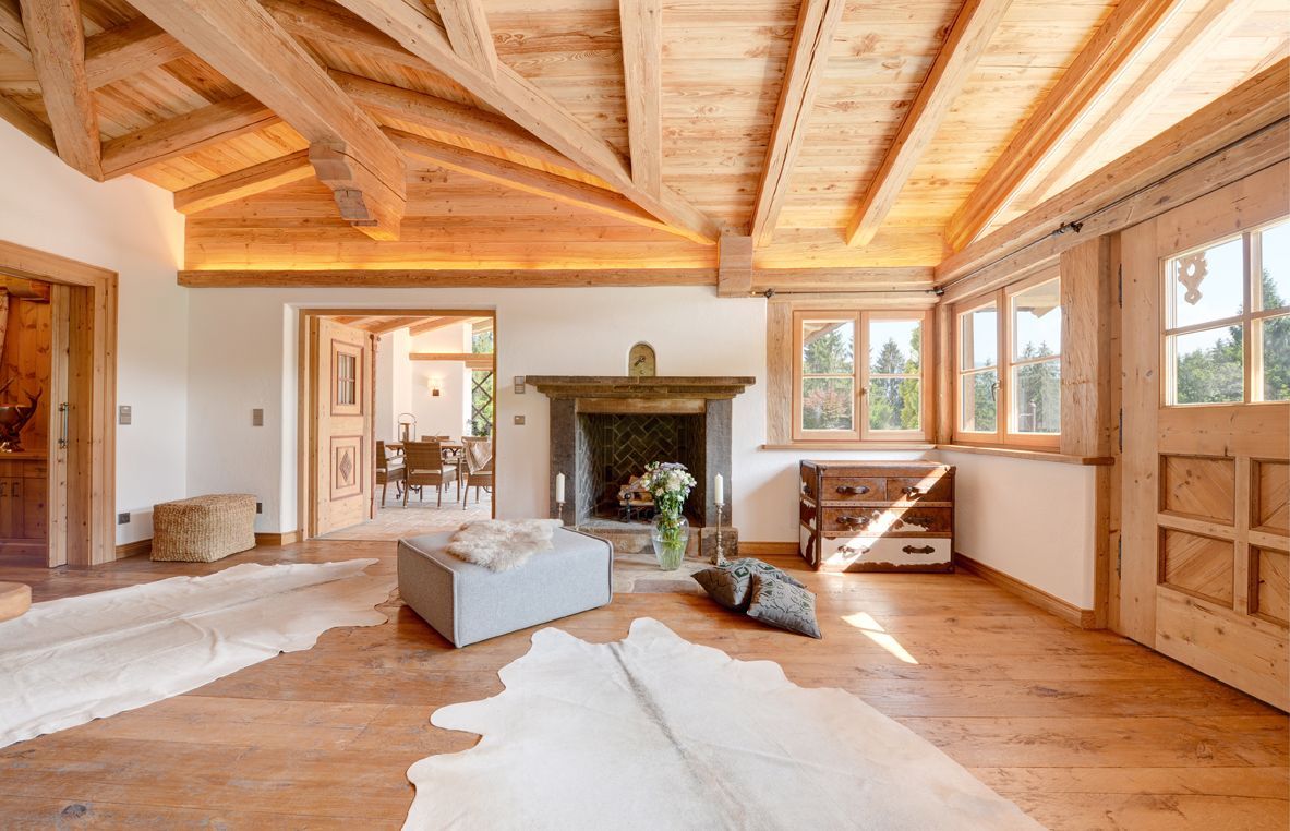 Wooden-beamed living room with a fireplace, cowhide rugs, and large windows. Sunlight streams in.