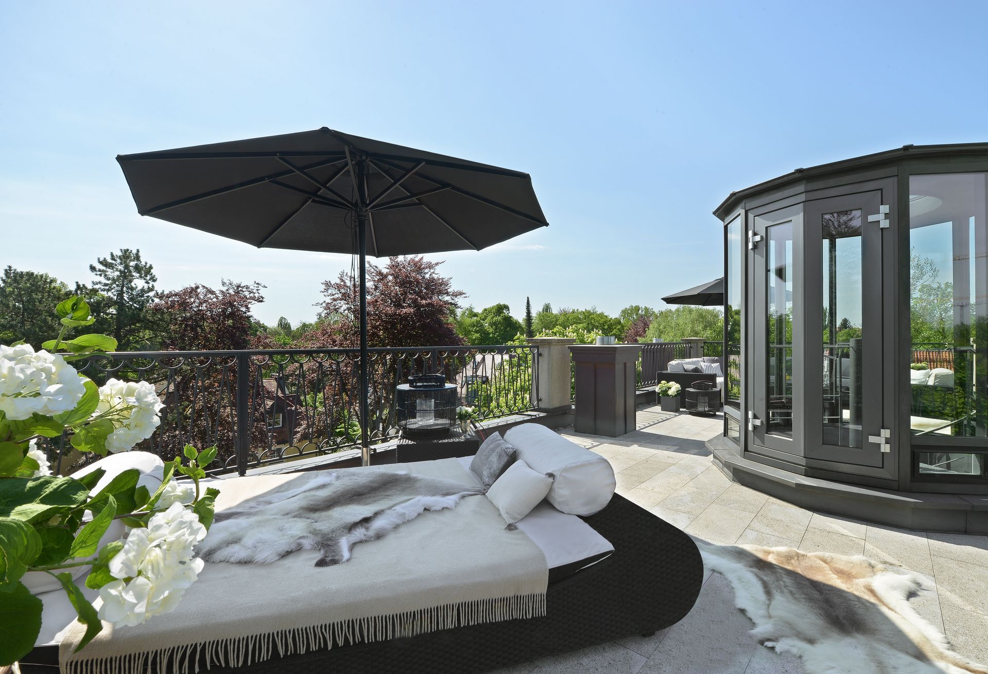 Rooftop patio with a daybed, umbrella, and a glass-enclosed structure. Sunny day.