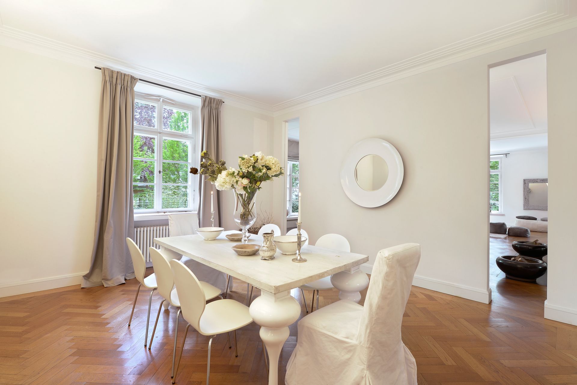 Dining room with white table, chairs, flowers, curtains, and wooden floor.