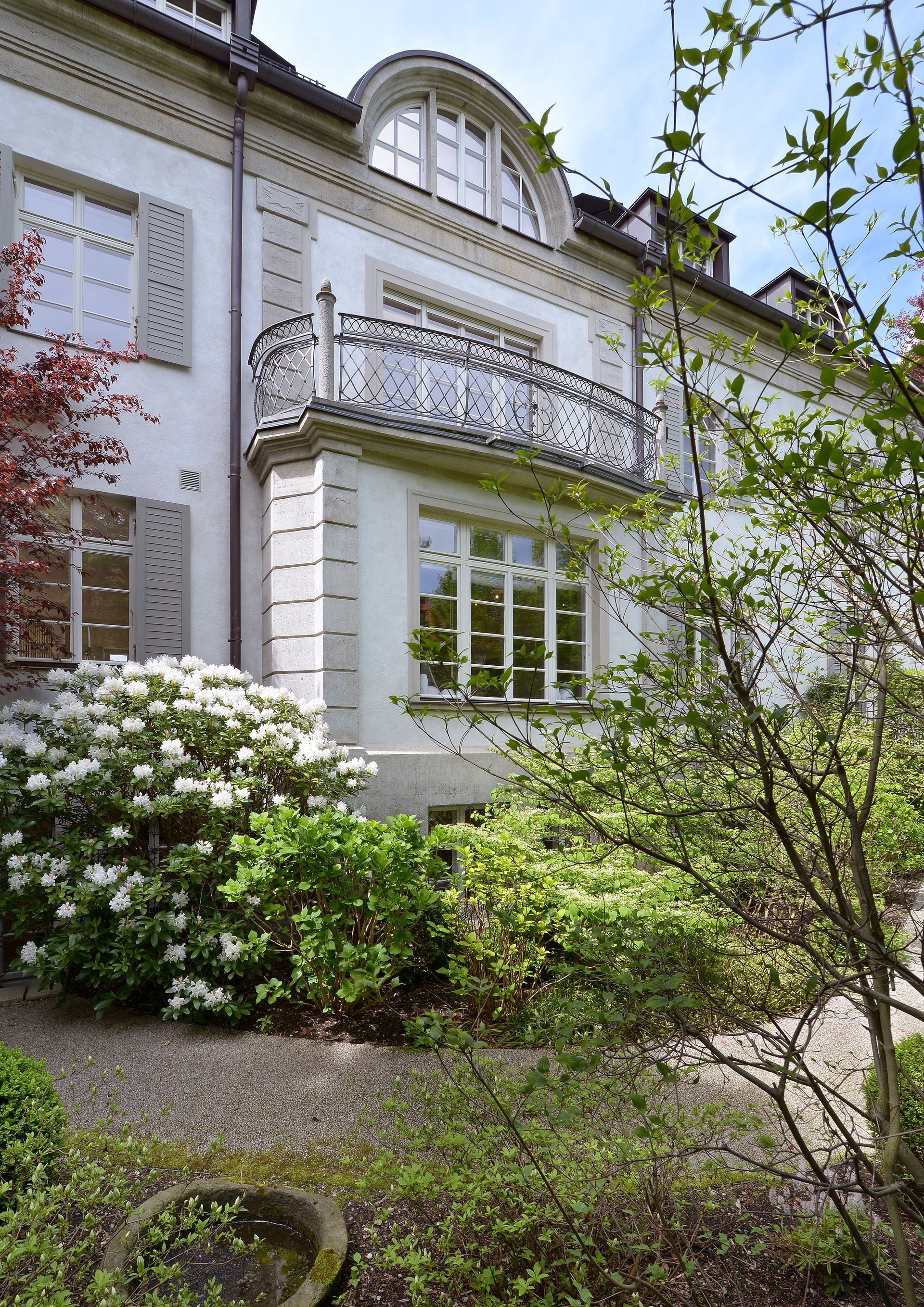White stucco house with balcony and shutters, surrounded by lush green and flowering bushes.