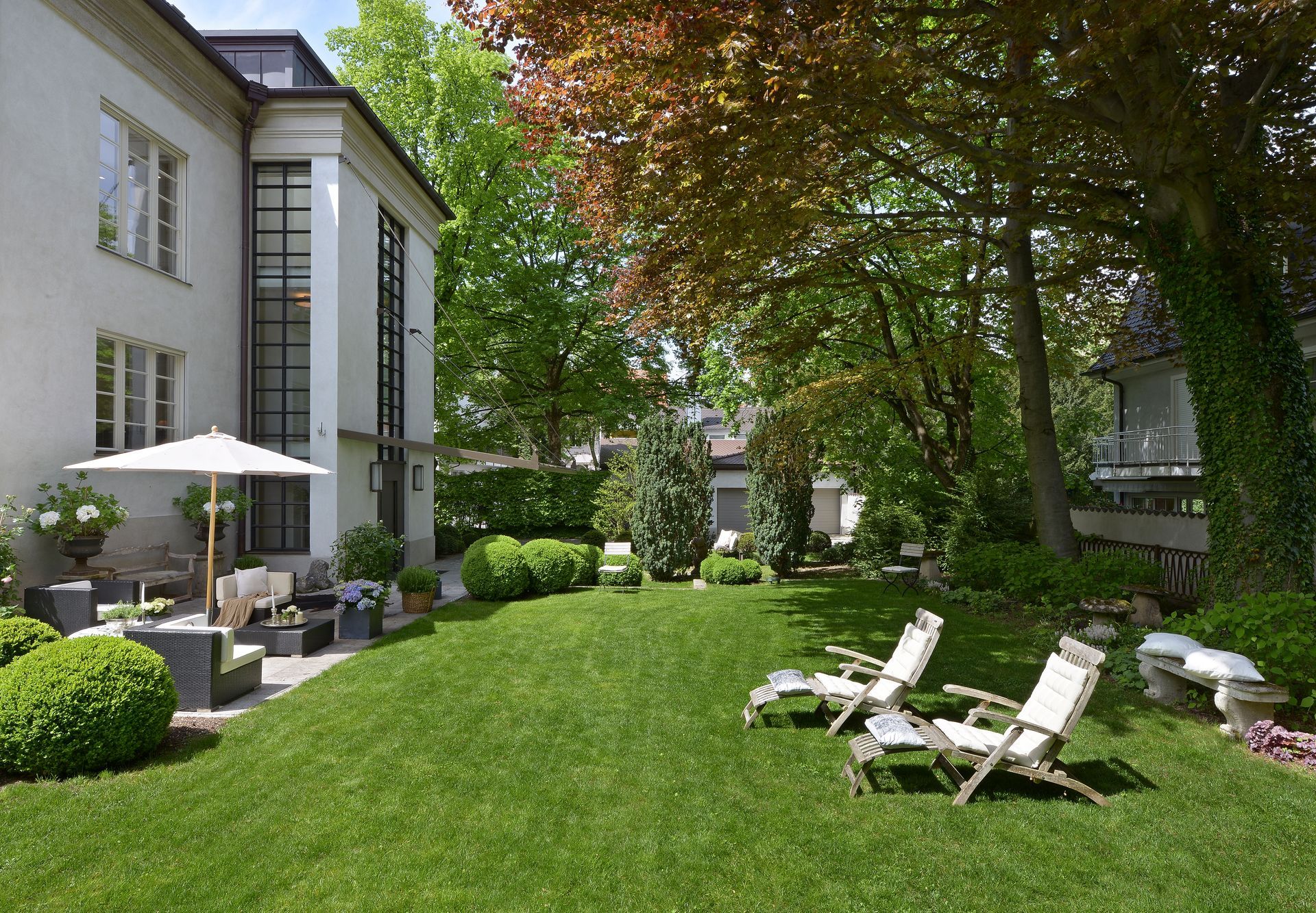Backyard with a lawn, trees, and lounge chairs next to a white house with a patio.